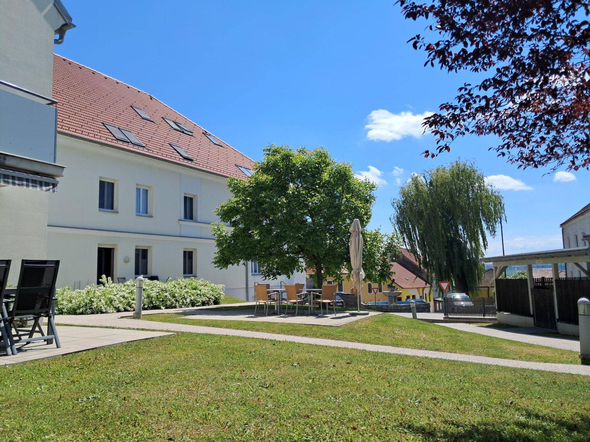 Sonniger Gartenbereich mit Sitzgelegenheiten und Blick auf den überdachten Parkplatz.