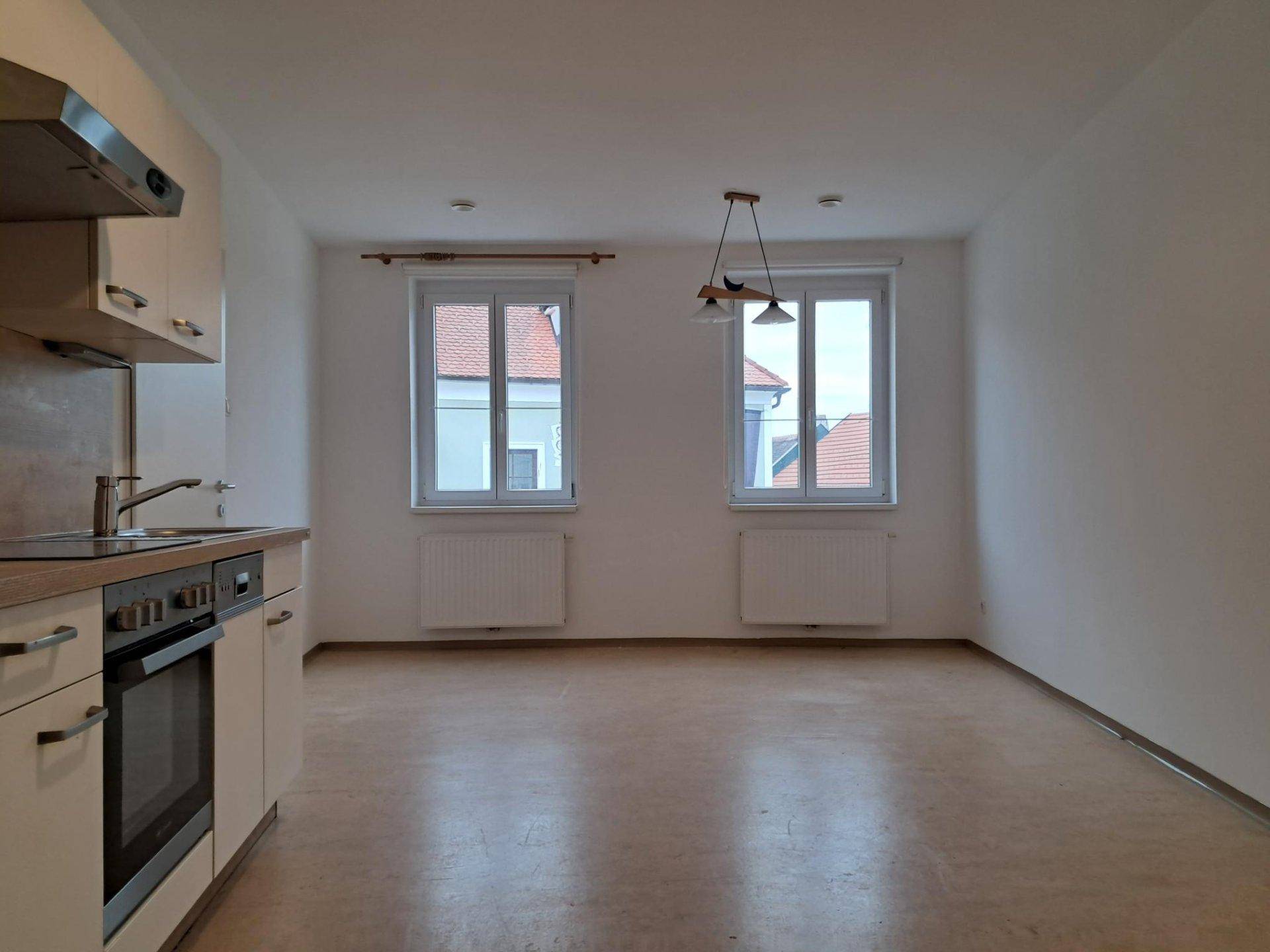 Heller Wohnraum mit integrierter Küchenzeile und zwei Fenstern mit Blick nach draußen.