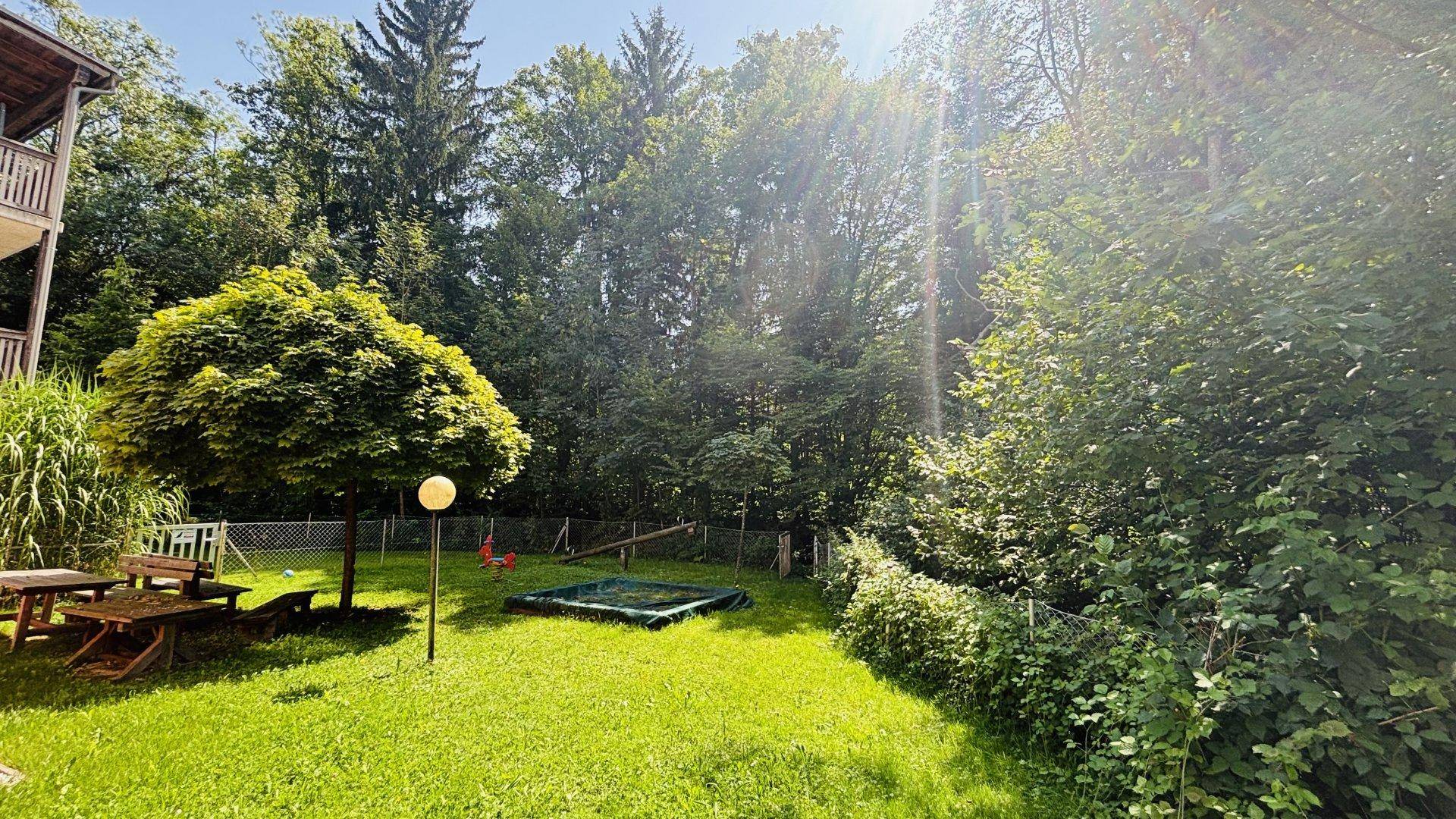 Sonniger Gemeinschaftsgarten mit Sandkasten, Schaukel und viel Grünfläche umgeben von Bäumen.