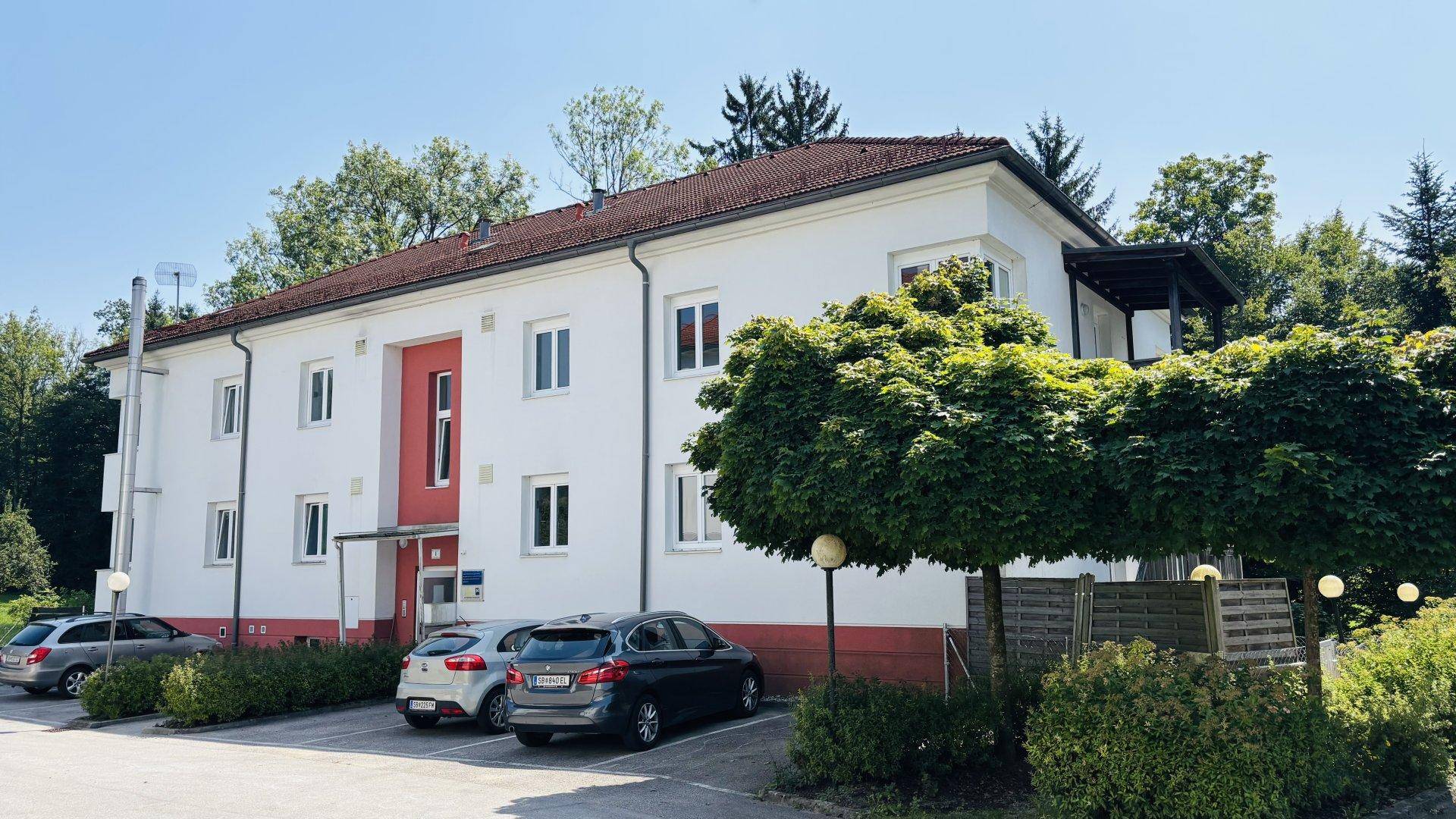 Modernes Mehrfamilienhaus mit weißer Fassade, roten Akzenten und Parkplätzen vor dem Gebäude.