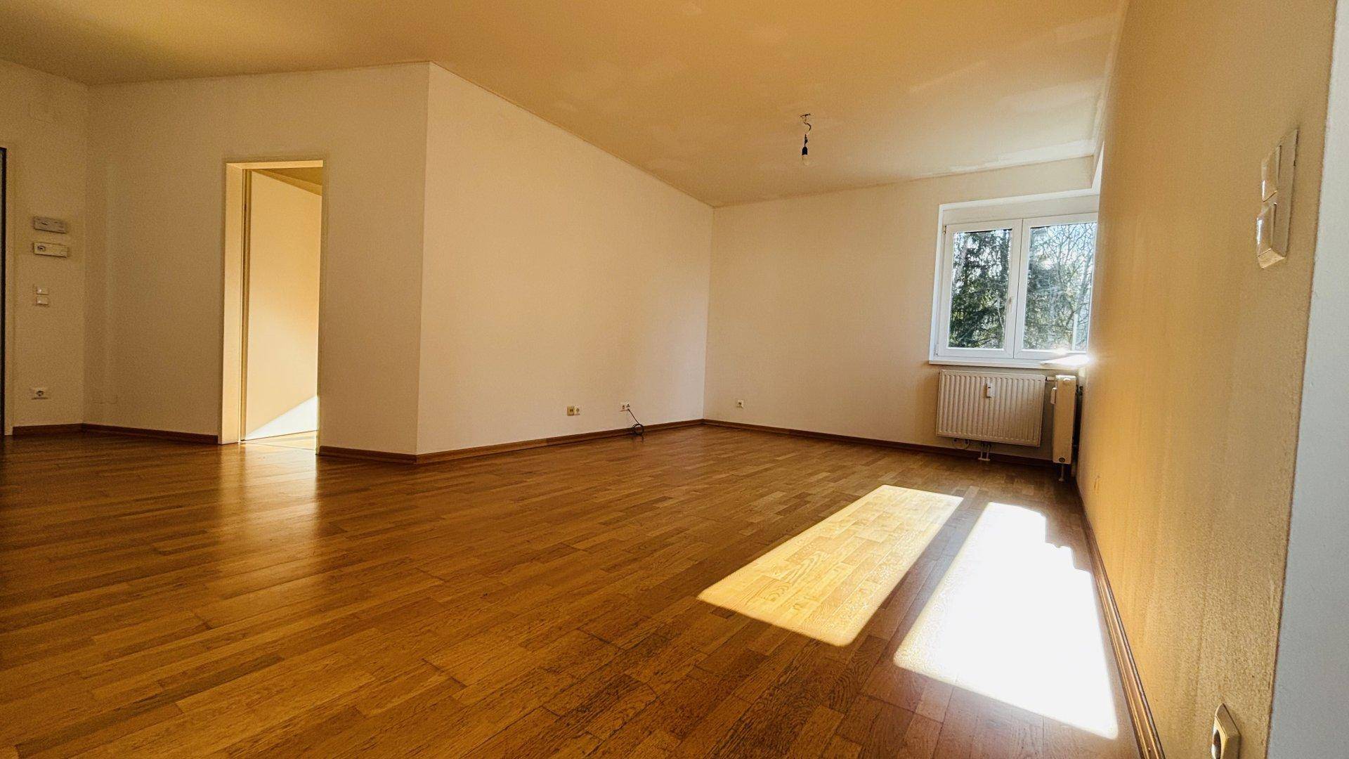 Großes Wohnzimmer mit Parkettboden, hellen Wänden und einem Fenster, das viel Licht hereinlässt.