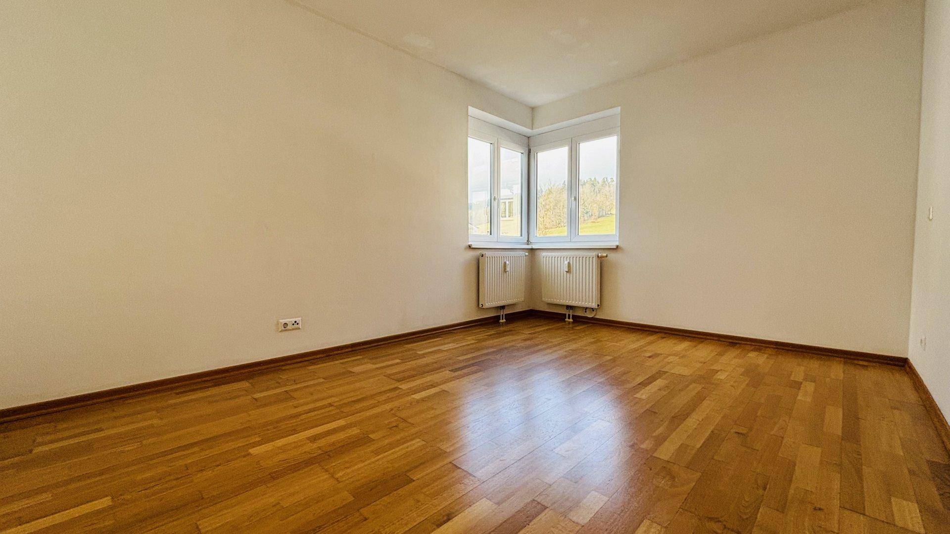 Helles Schlafzimmer mit Parkettboden, großen Fenstern und Heizkörpern unter dem Fenster.