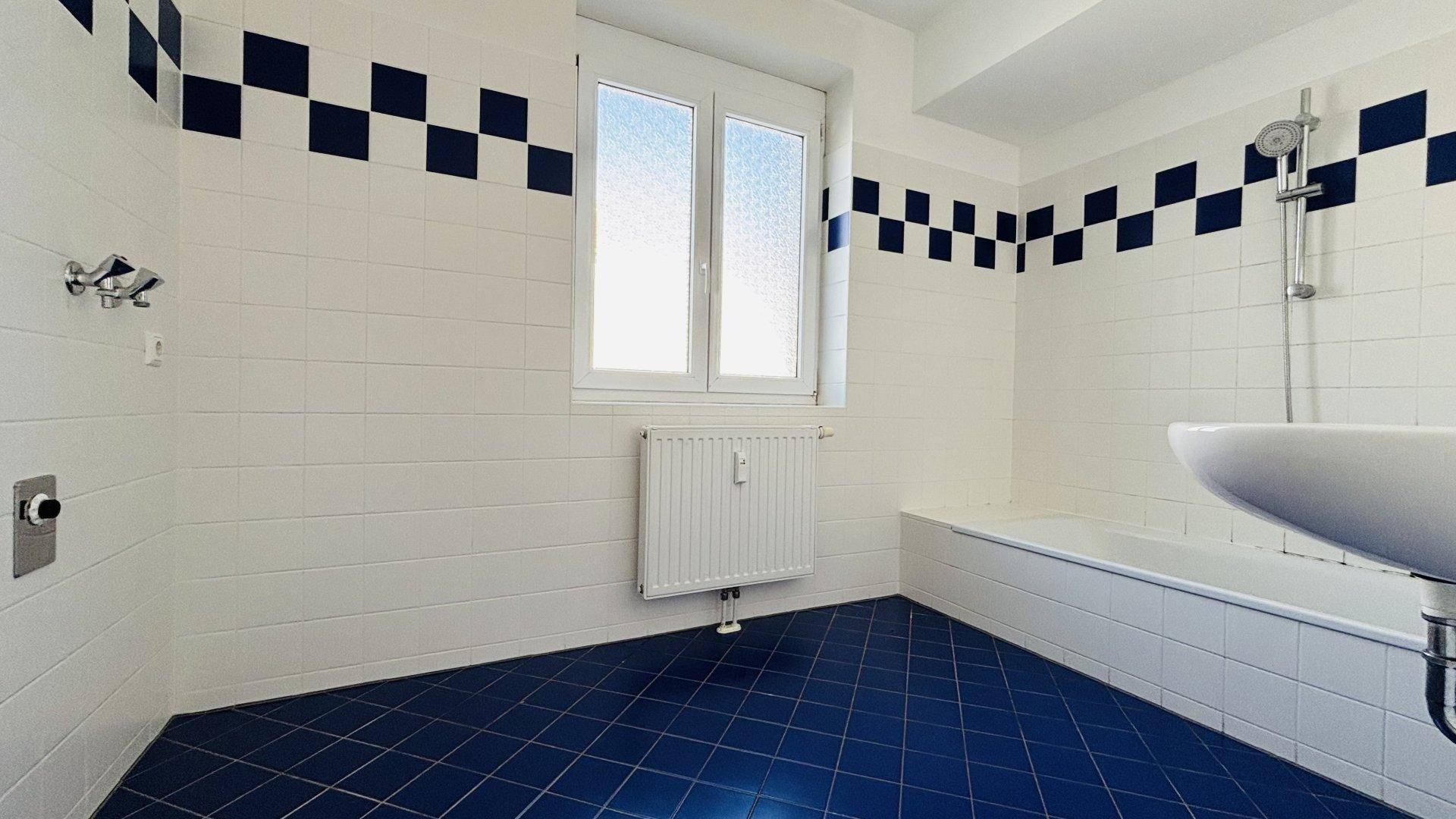 Badezimmer mit blauen und weißen Fliesen, Badewanne mit Duschfunktion und Fenster.