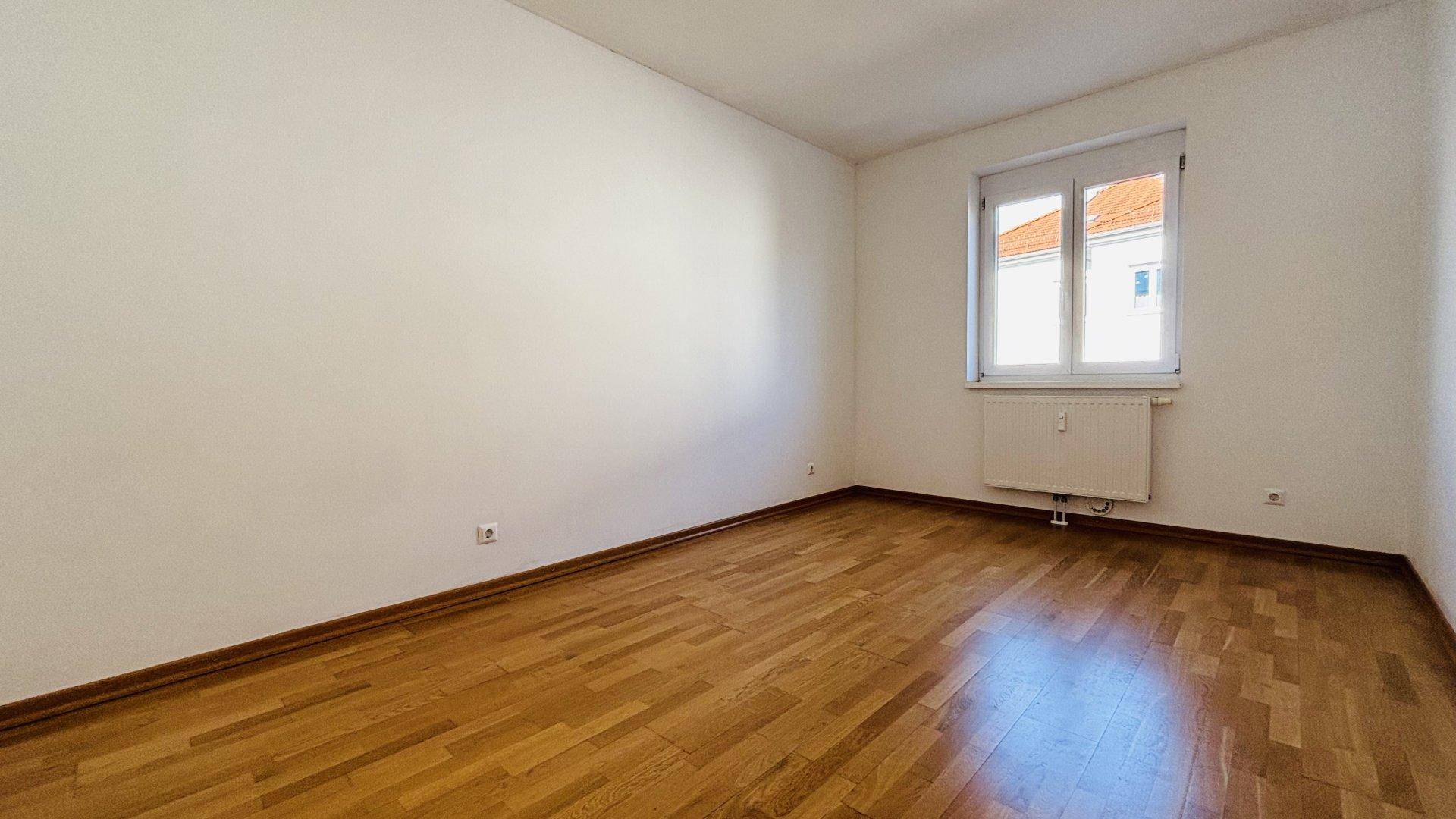 Schlafzimmer mit Parkettboden, weiß gestrichenen Wänden und einem Fenster mit Blick nach draußen.