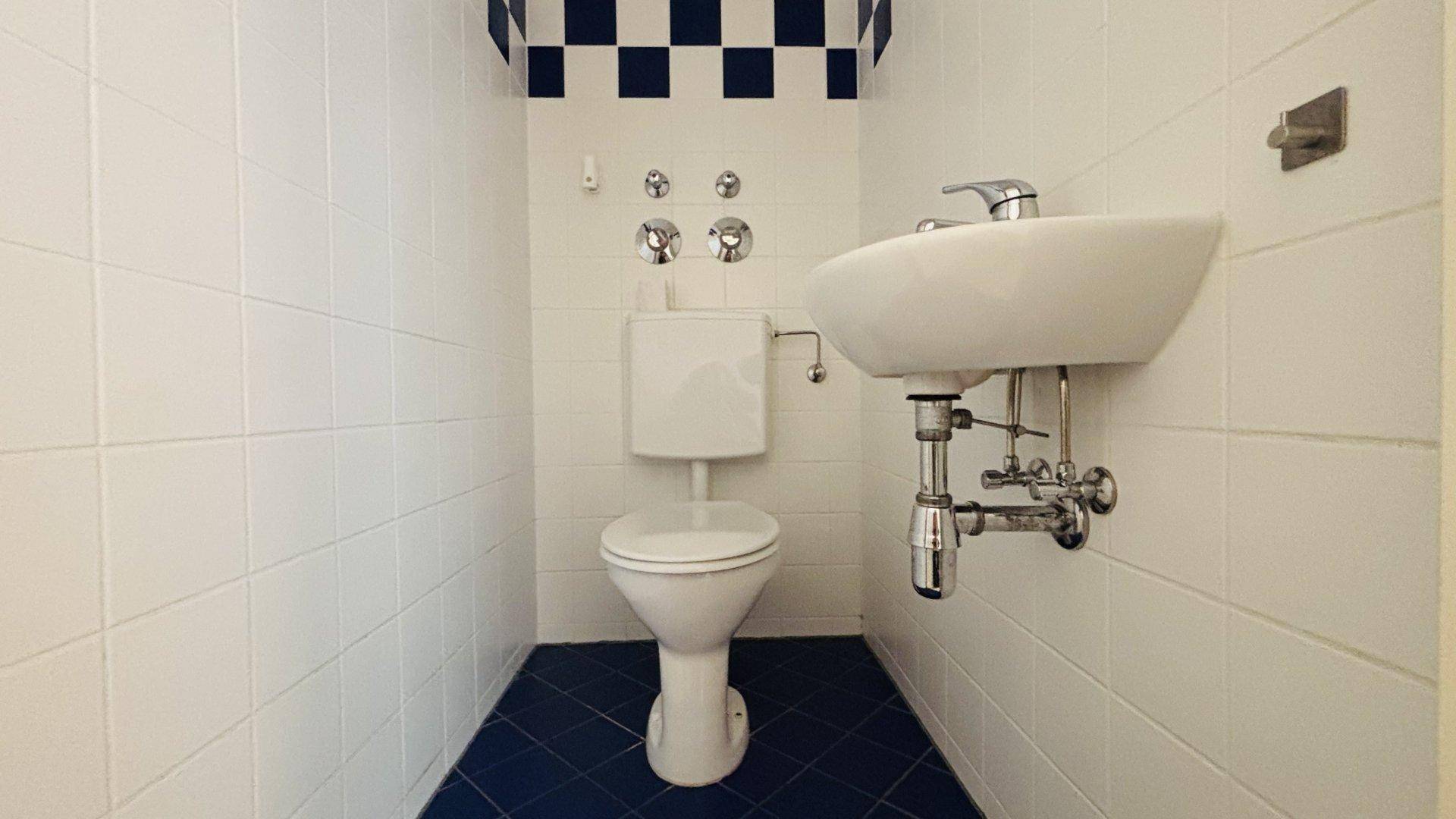 Separate Toilette mit Waschbecken, weißen Wandfliesen und einem blauen Fliesenboden.
