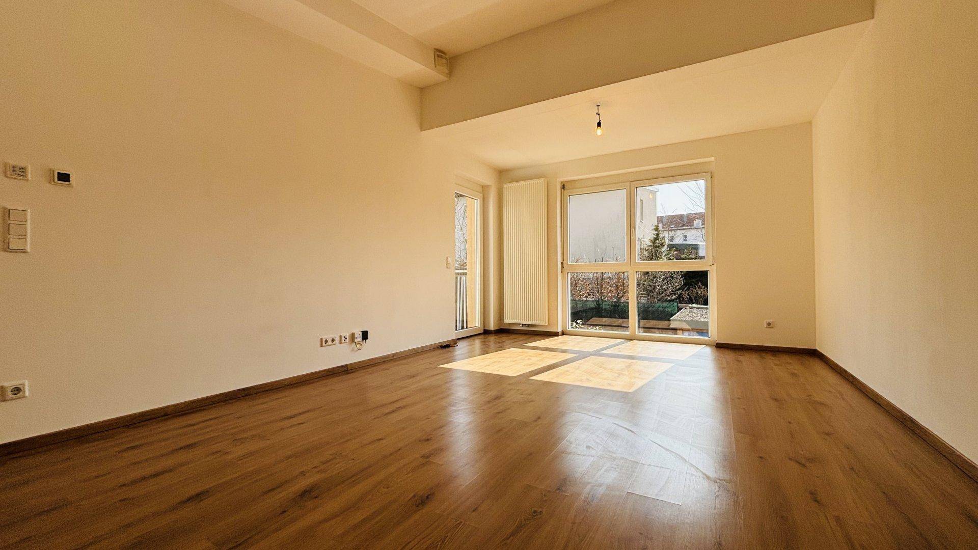 Helles Wohnzimmer mit Laminatboden, großen Fenstern und Zugang zu einem kleinen Balkon.