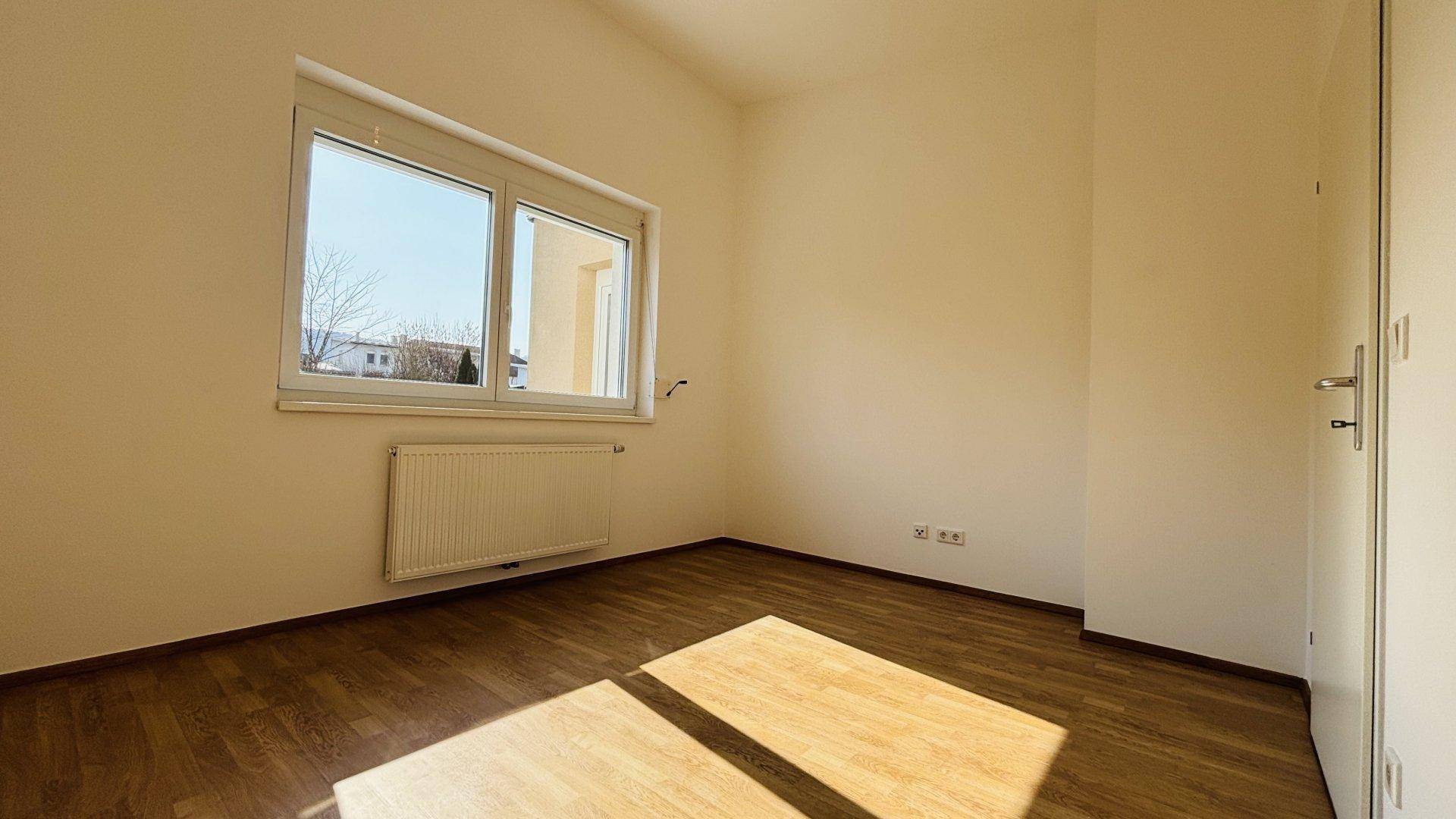 Helles Schlafzimmer mit Laminatboden und großem Fenster, das viel Tageslicht hereinlässt.