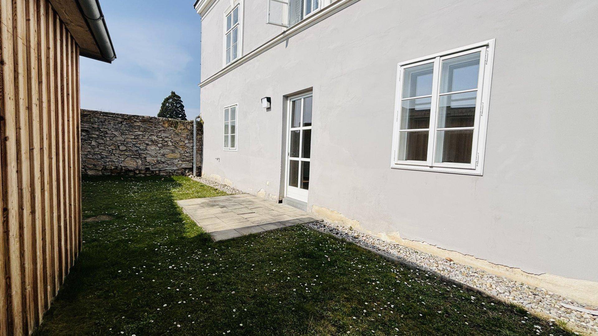 Kleiner Gartenbereich mit gepflasterter Terrasse und Zugang zum Gebäude, umgeben von Grün.