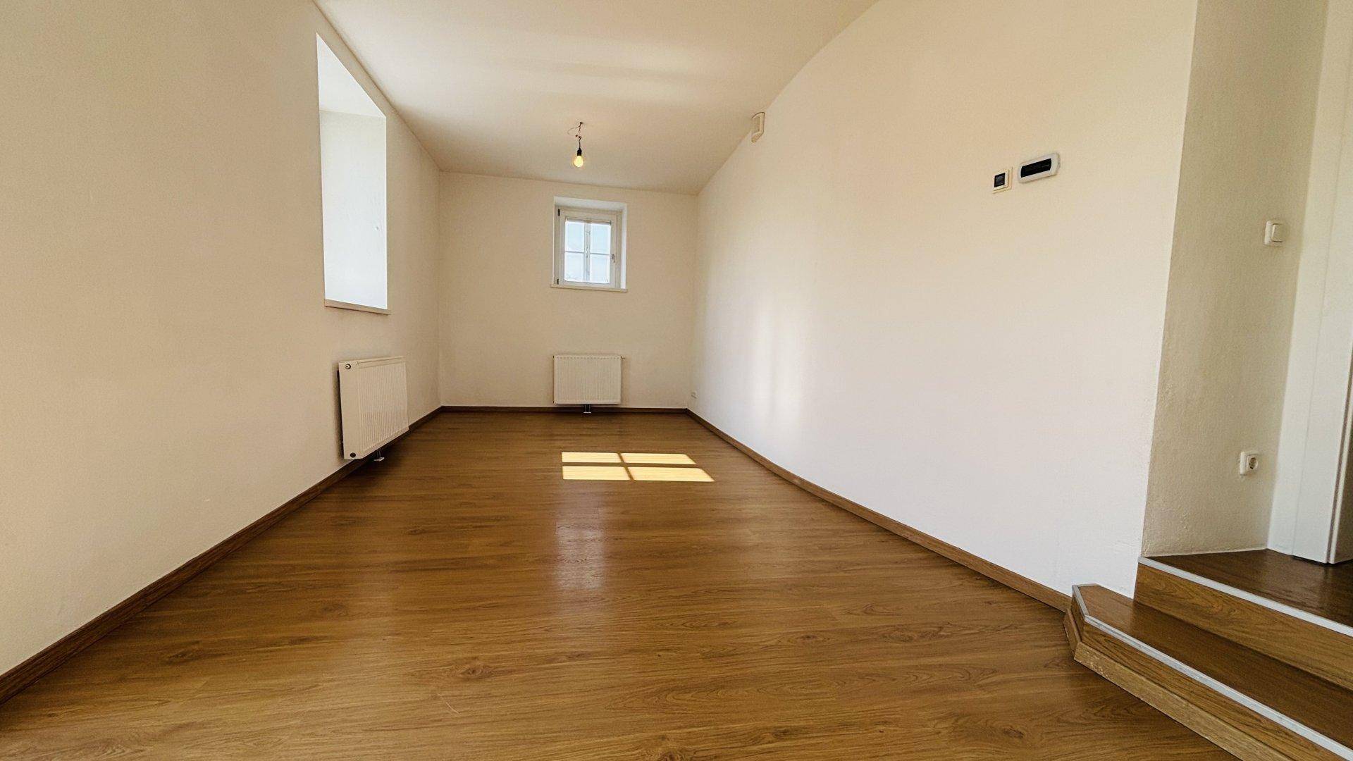 Lichtdurchfluteter Raum mit Holzboden und einem kleinen Treppenaufgang, ideal für flexibles Wohnen.