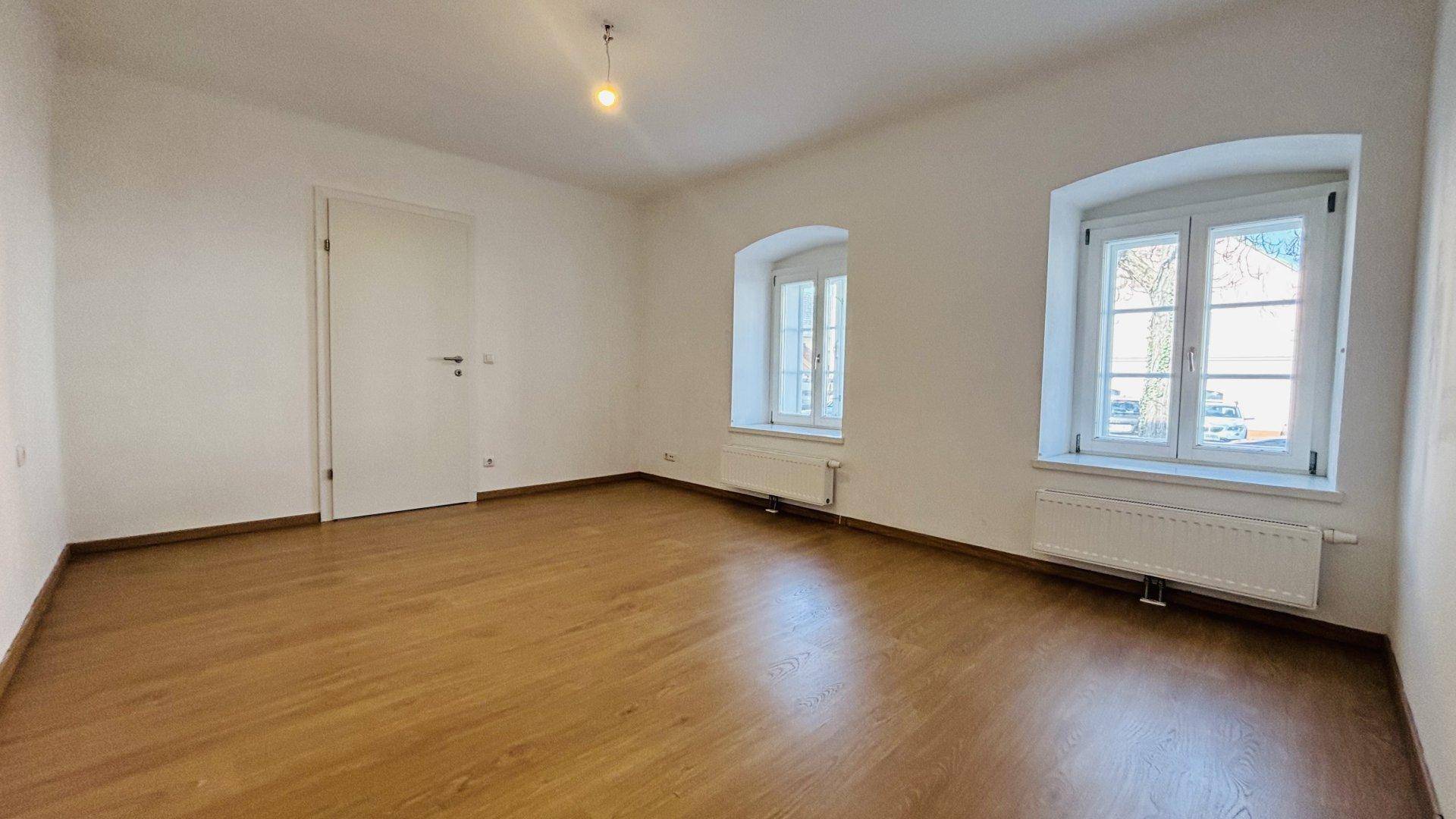 Großzügiger Wohnraum mit Holzboden und zwei Fenstern, die viel Tageslicht hereinlassen.