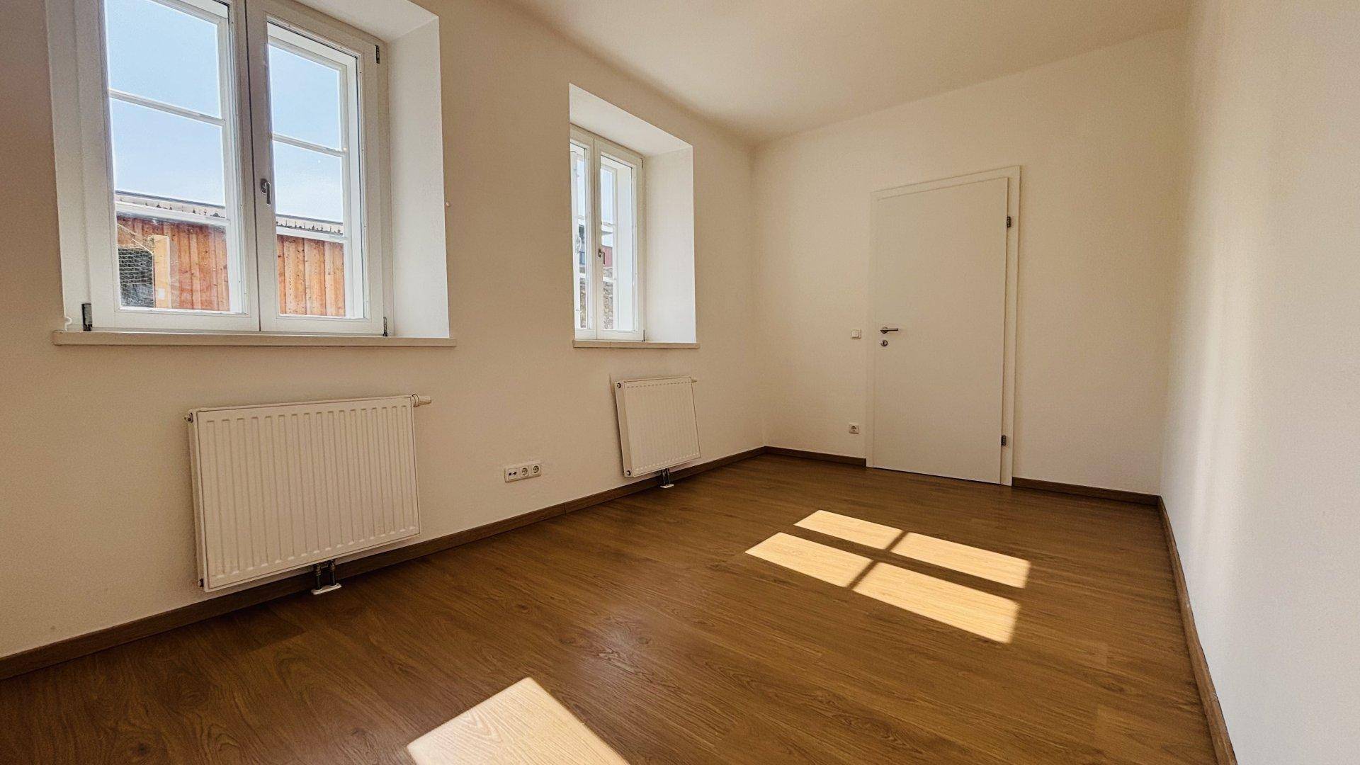 Sonniges Zimmer mit Holzboden und zwei Fenstern, ideal als Schlaf- oder Arbeitszimmer nutzbar.