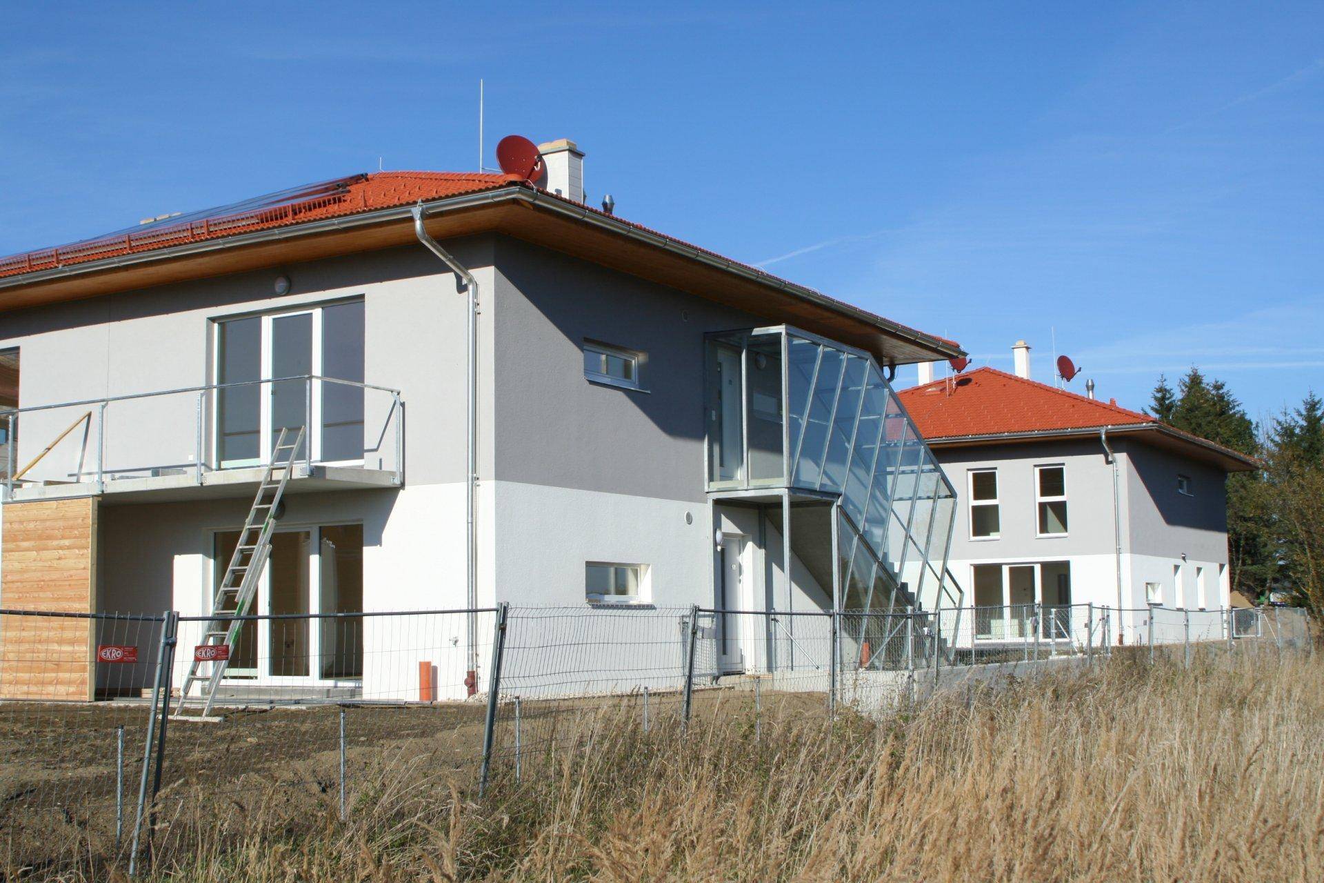 Zwei moderne Neubauten mit rotem Ziegeldach und einer verglasten Außentreppe in sonniger Umgebung.