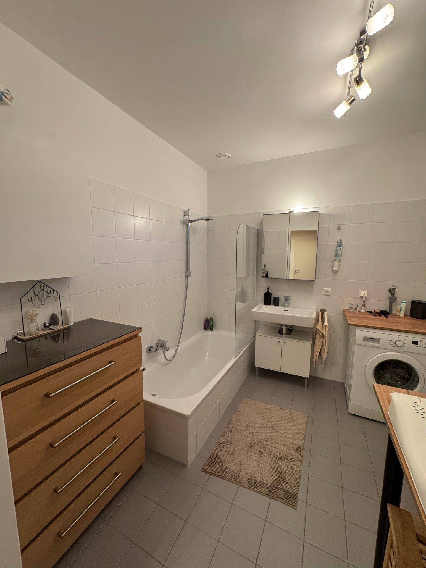 Funktionales Badezimmer mit Badewanne, Waschbecken, Waschmaschine und hellen Fliesen.