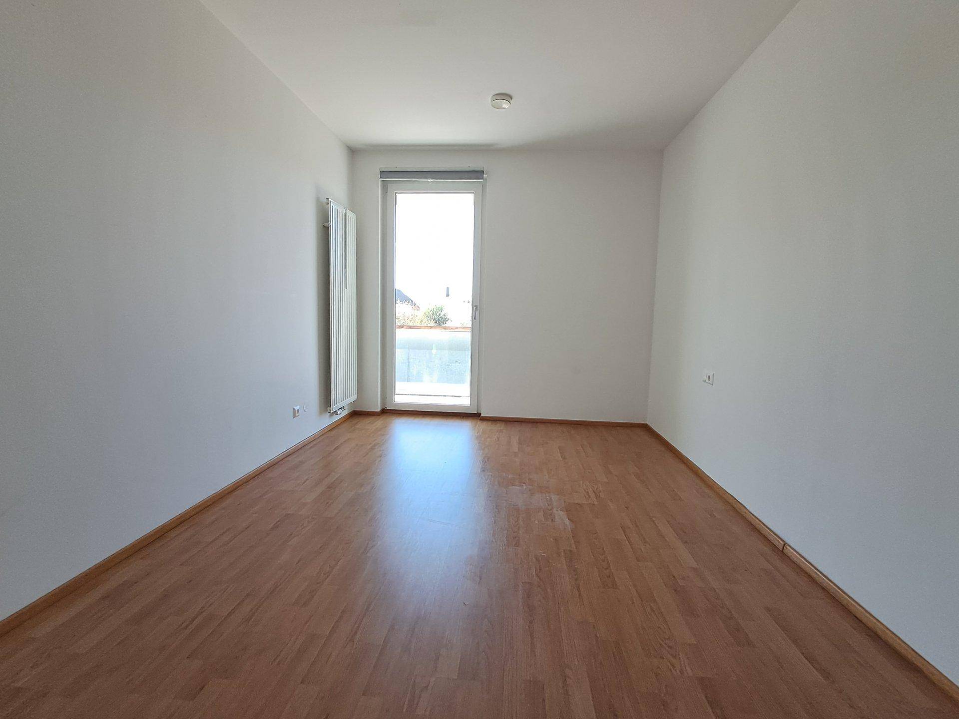 Helles, leeres Zimmer mit Laminatboden und Zugang zu einem Balkon.