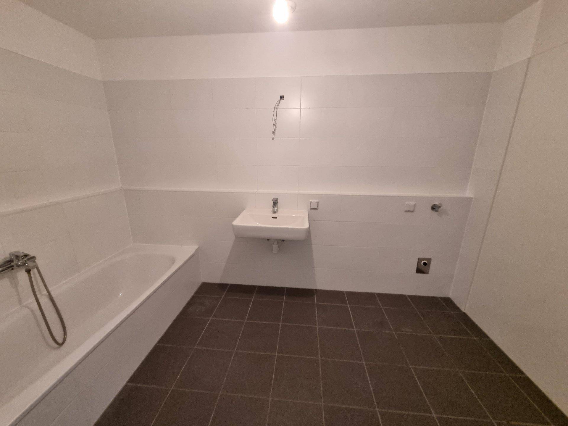 Leeres Badezimmer mit Badewanne, Waschbecken und dunklen Bodenfliesen, bereit zur Einrichtung.