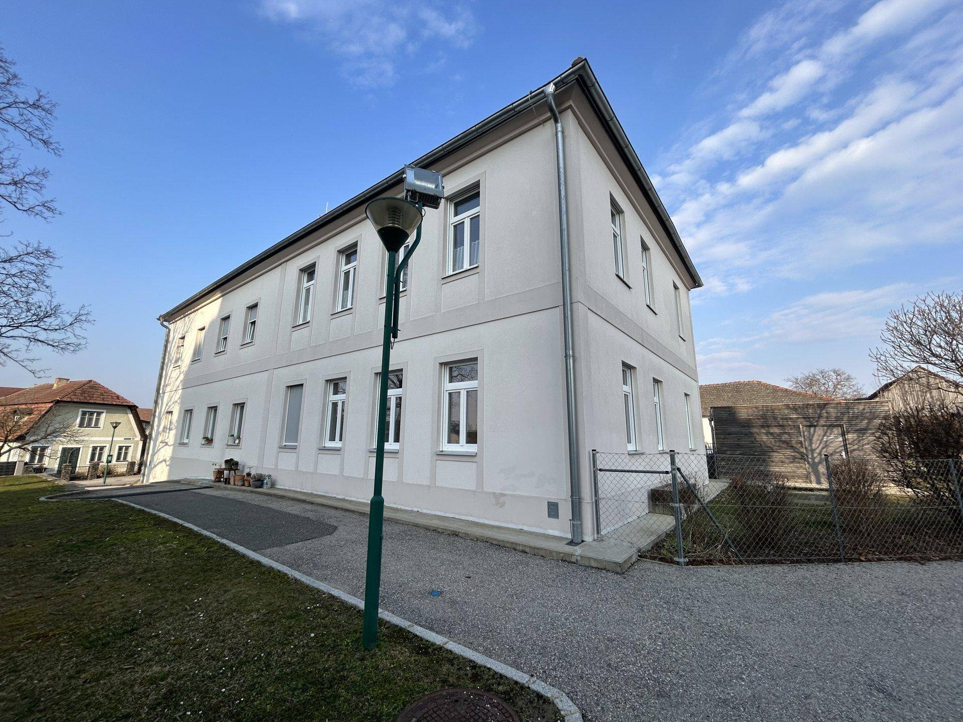 Seitenansicht eines hellen Gebäudes mit vielen Fenstern und einem Kiesweg.