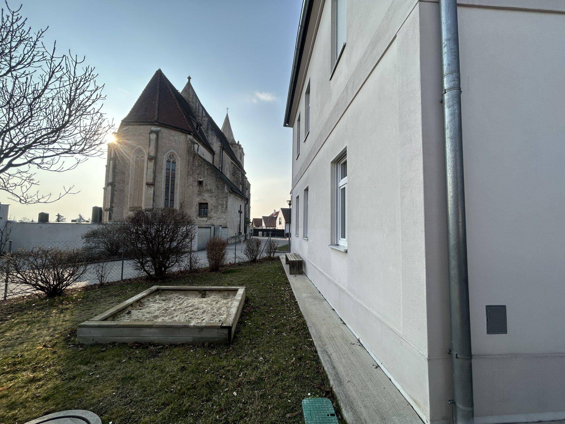 Außenbereich mit Sandkasten und Blick auf eine historische Kirche im Hintergrund.