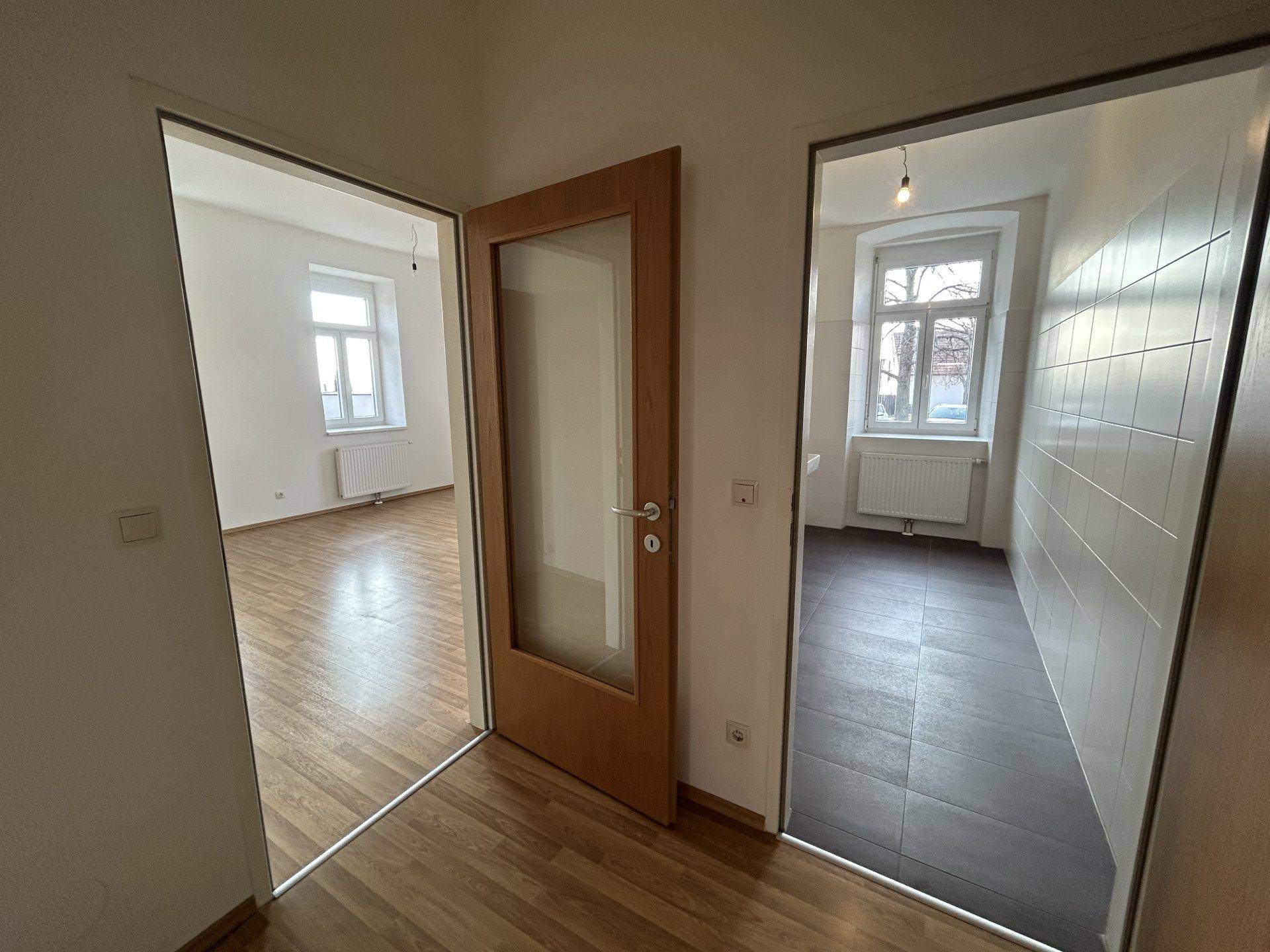 Flur mit Holztür und Zugang zu zwei weiteren hellen Räumen mit unterschiedlichen Böden.