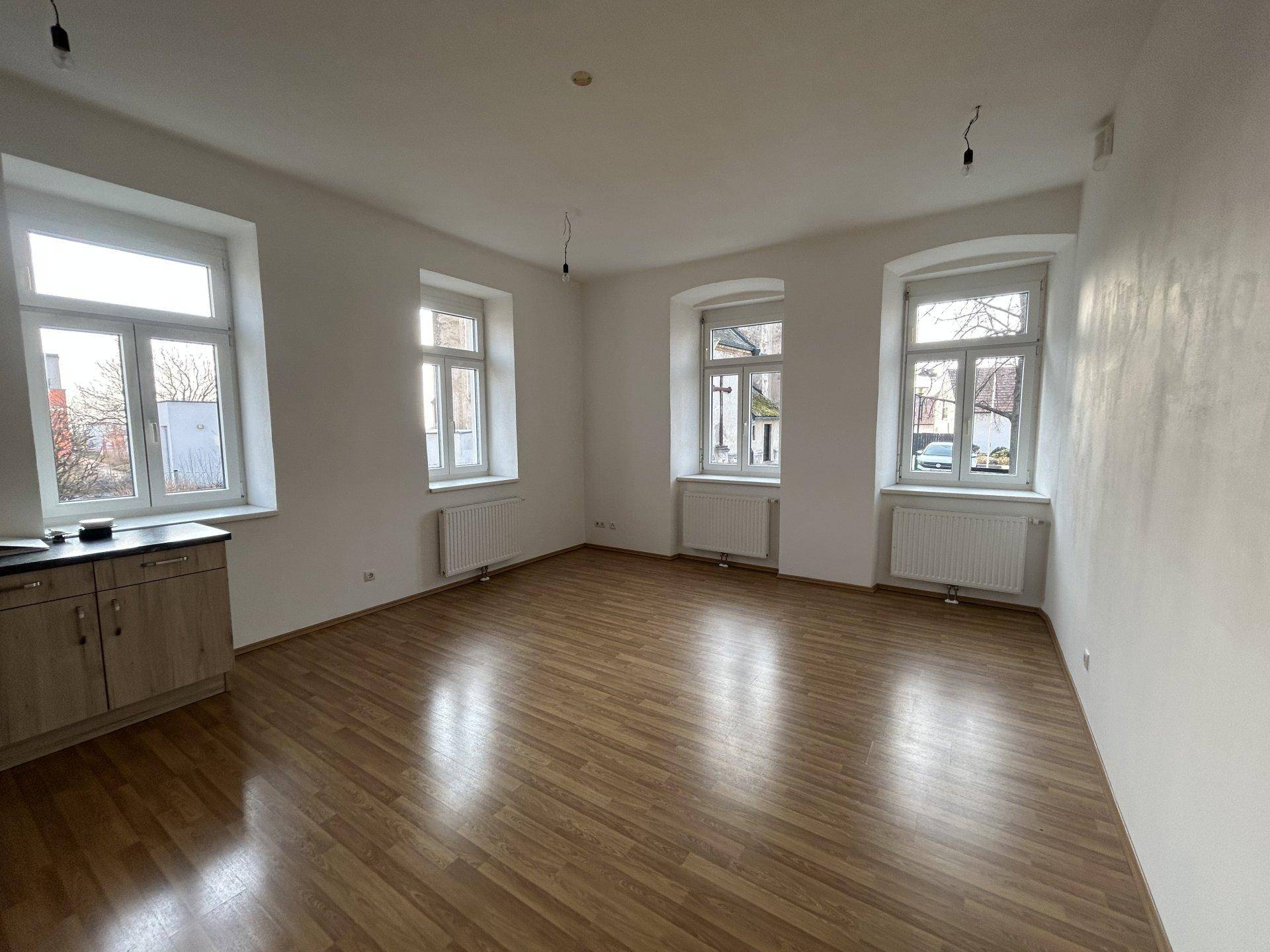 Heller Wohnraum mit drei Fenstern, Laminatboden und weiß gestrichenen Wänden.
