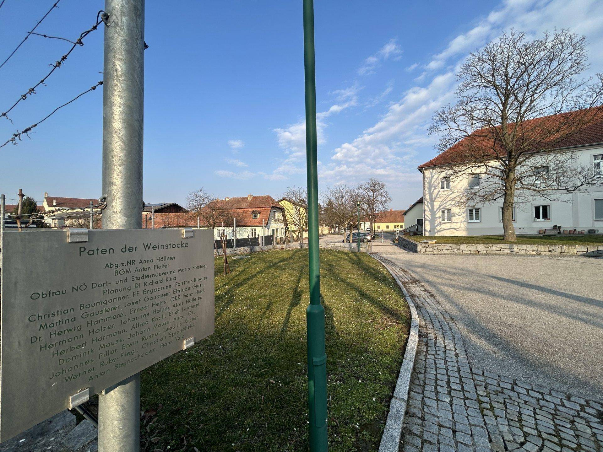 Ländliche Umgebung mit Kopfsteinpflasterweg, Grünflächen und einem Informationsschild.
