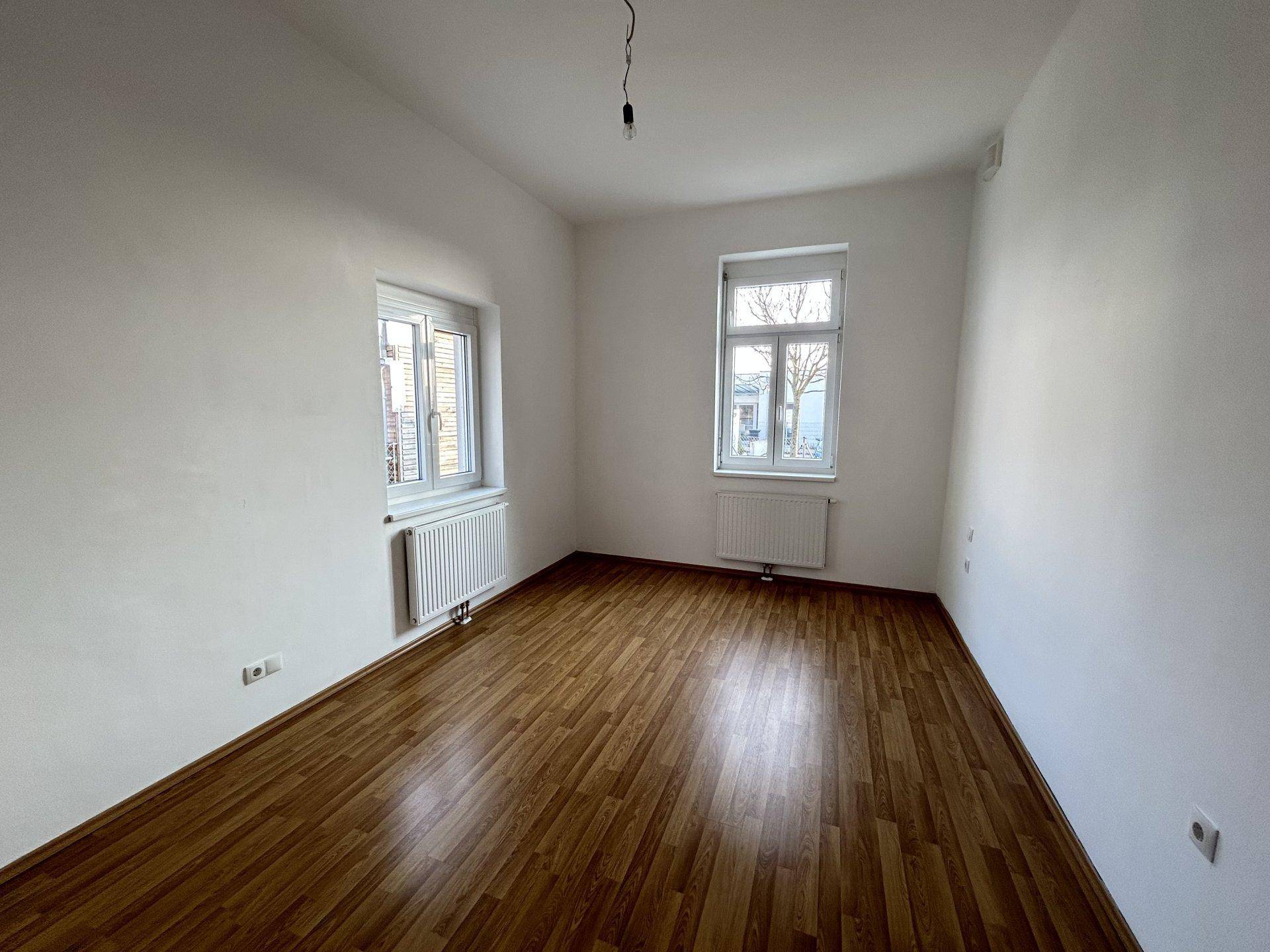 Schlafzimmer mit Laminatboden, zwei Fenstern und weiß gestrichenen Wänden.
