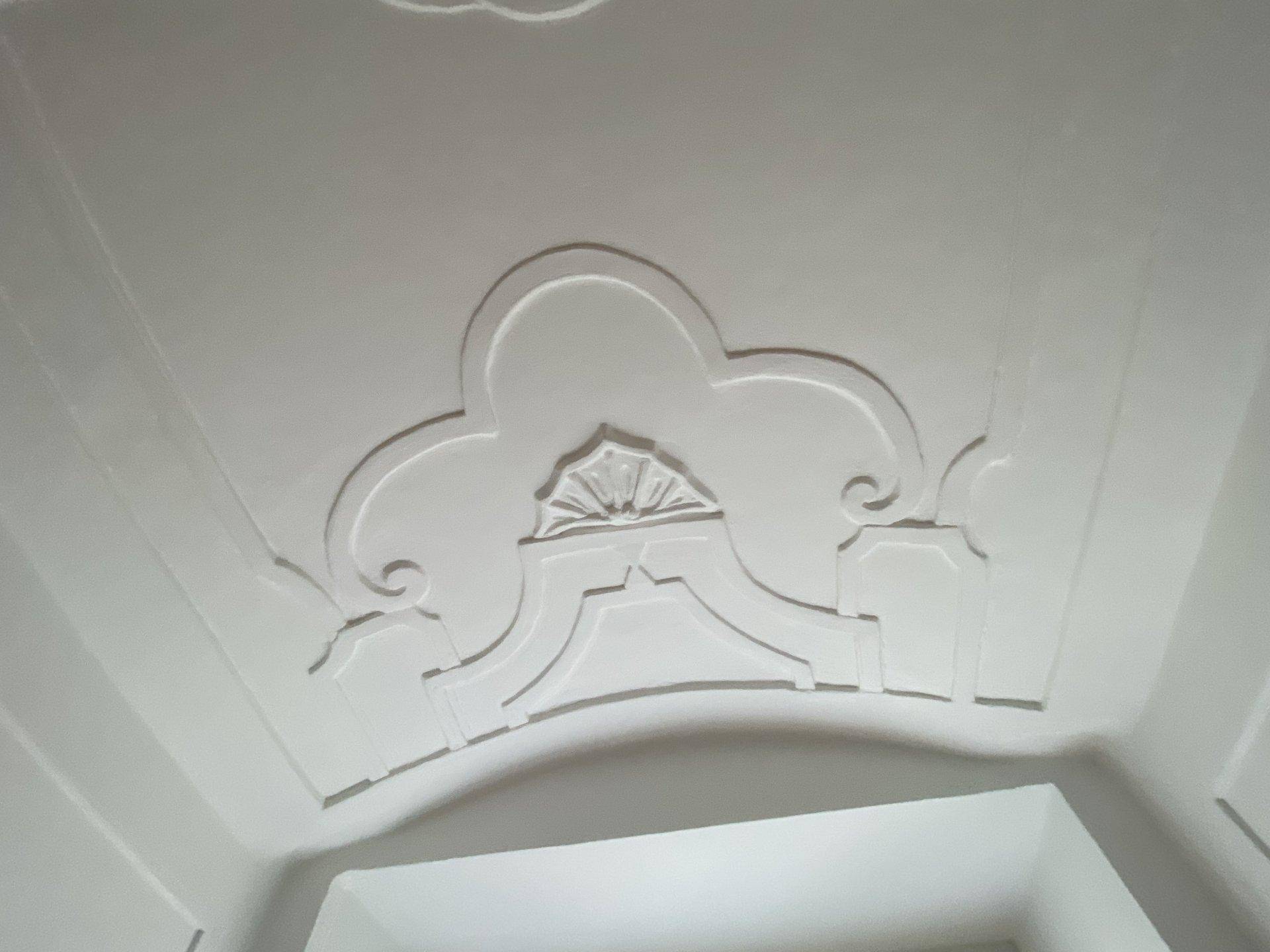 Detailaufnahme einer weißen Decke mit kunstvollen Stuckelementen und geschwungenen Formen, die den Raum schmücken.