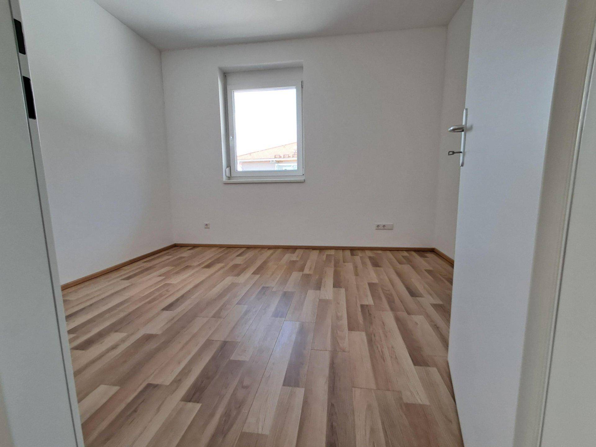 Helles Schlafzimmer mit Laminatboden und einem Fenster mit Blick nach draußen.