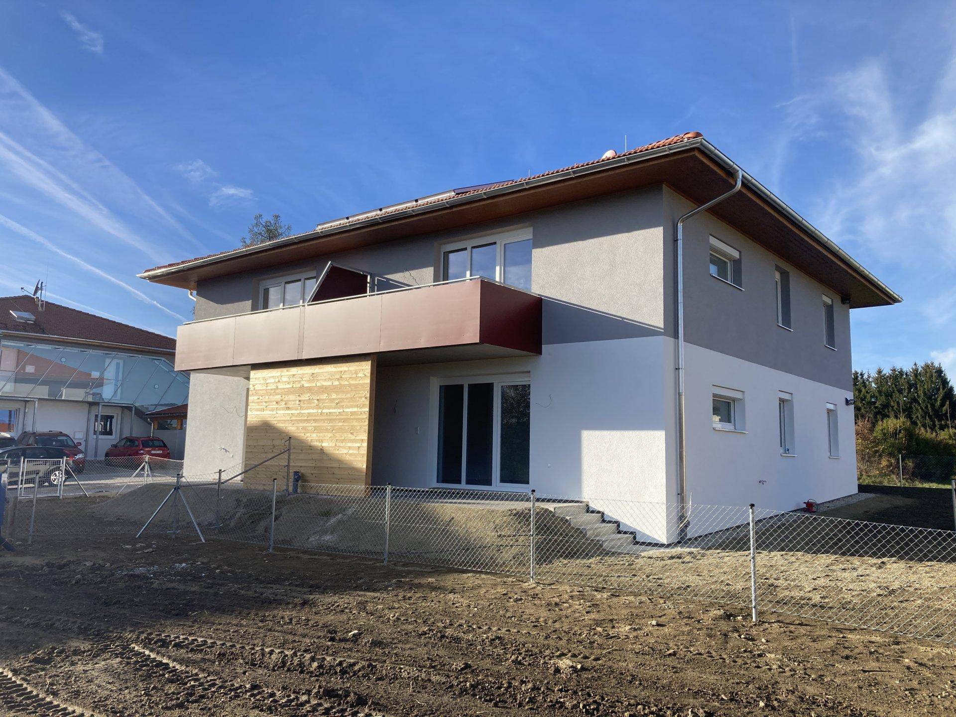 Seitenansicht eines modernen Neubaus mit rotem Balkon und vorbereitetem Gartenbereich.