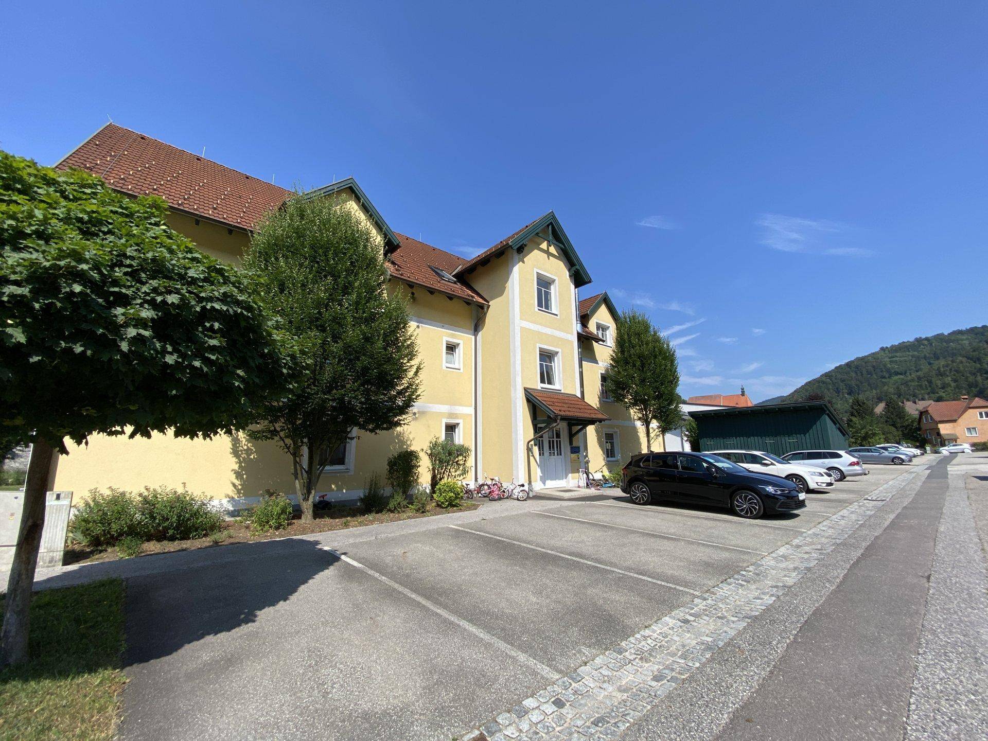 Vorderansicht des gelben Gebäudes mit Eingangsbereich und einem Parkplatz voller Autos unter blauem Himmel.