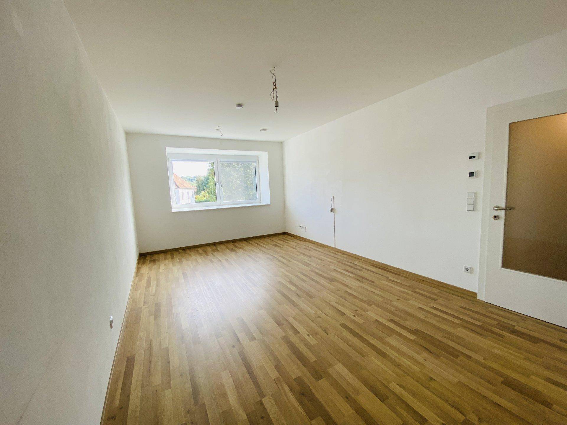 Helles, leeres Zimmer mit Holzparkettboden und einem Fenster mit Blick ins Grüne.