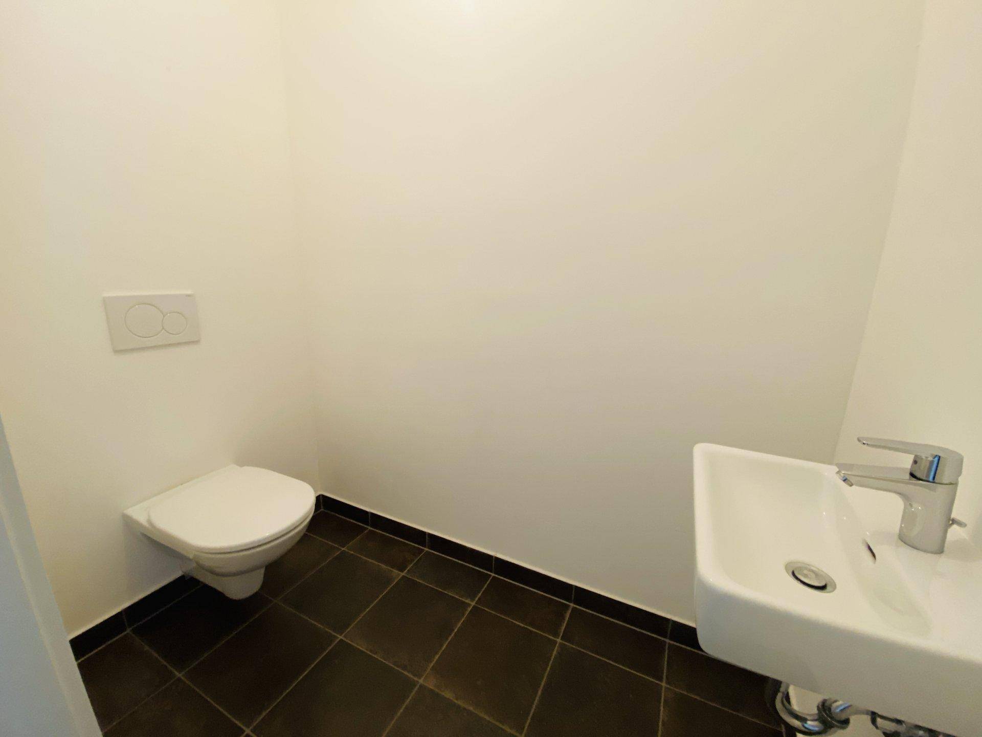 Separates WC mit wandhängender Toilette, kleinem Waschbecken und dunklen Bodenfliesen.