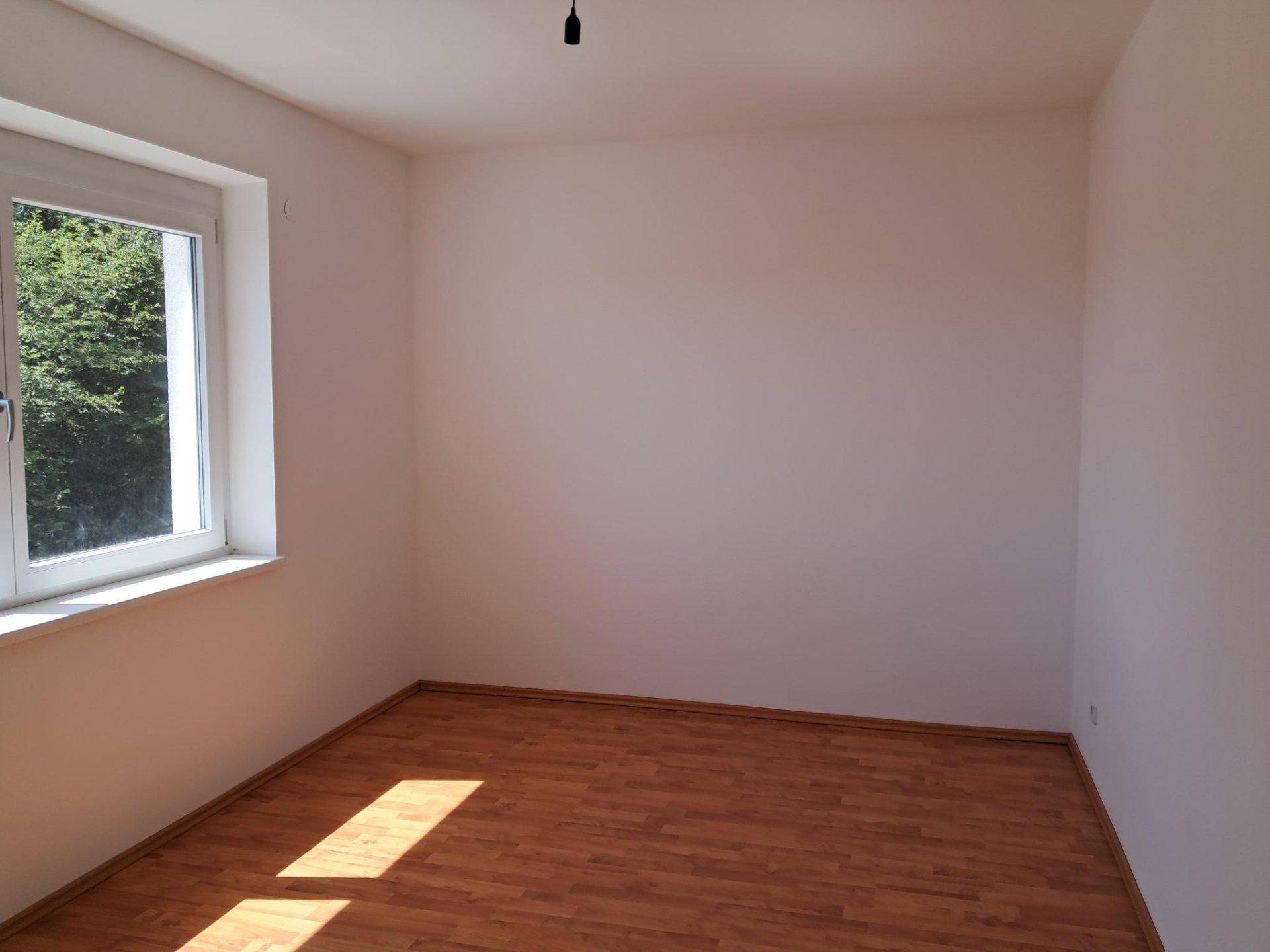 Sonniger Raum mit Laminatboden und einem großen Fenster, das viel Tageslicht hereinlässt.