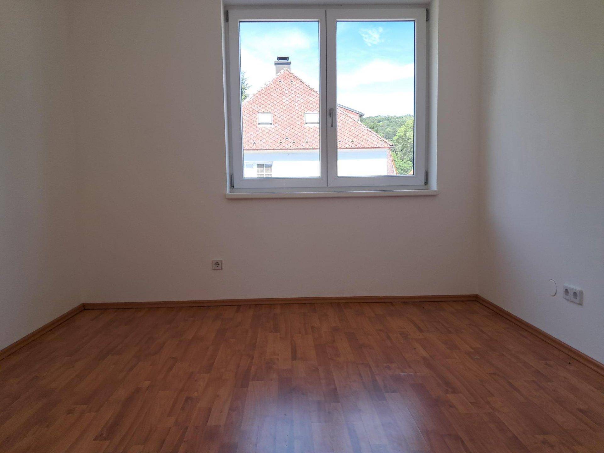 Helles Schlafzimmer mit Laminatboden und Blick auf ein gegenüberliegendes Ziegeldach.