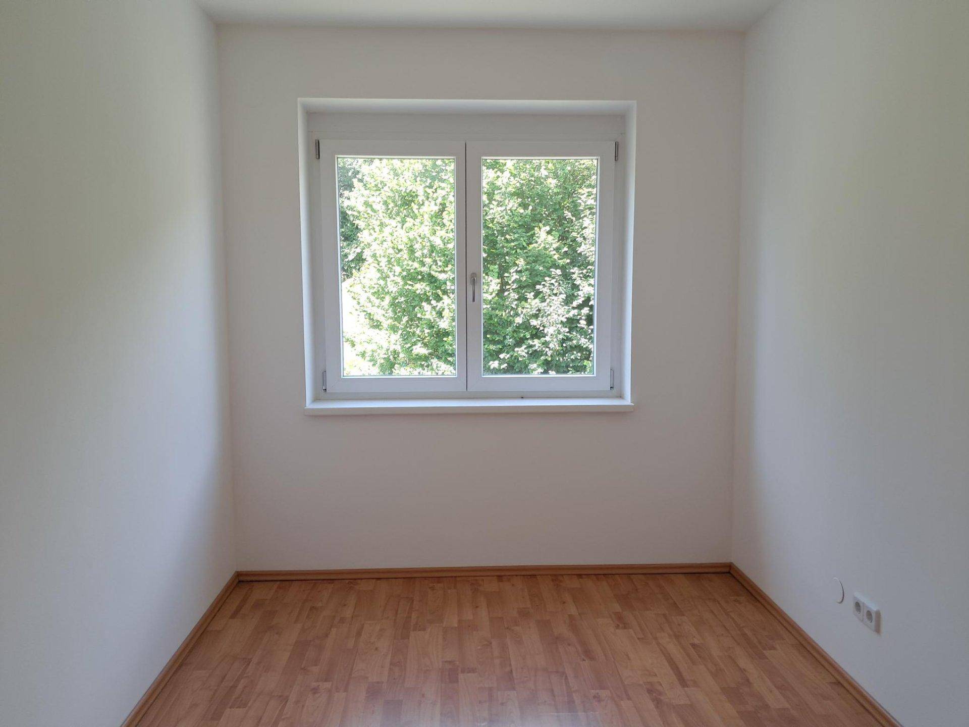 Freundliches Zimmer mit Laminatboden und Blick ins Grüne durch das große Fenster.