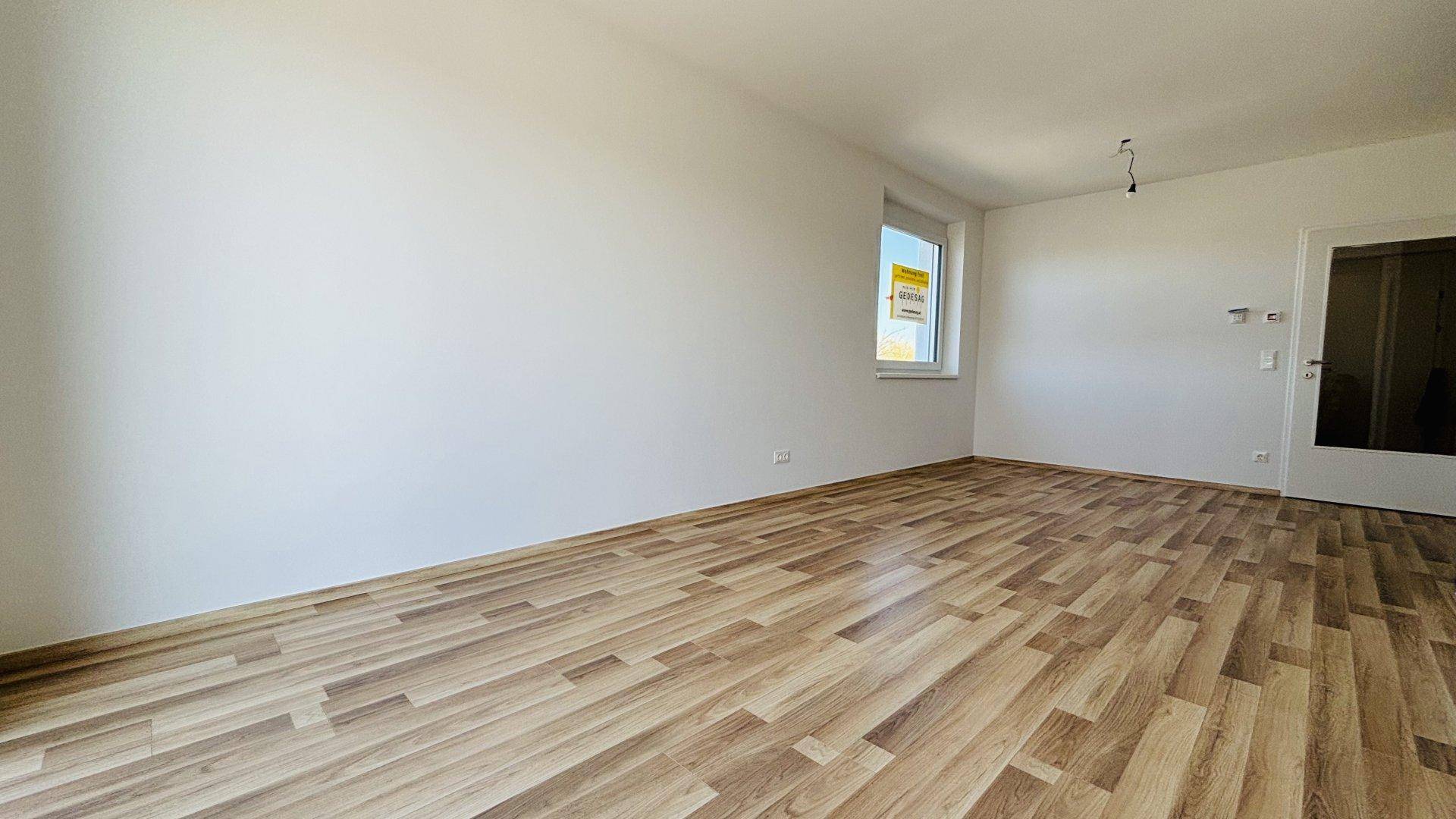 Leerer, heller Raum mit Laminatboden und einem großen Fenster, das viel Tageslicht hereinlässt.