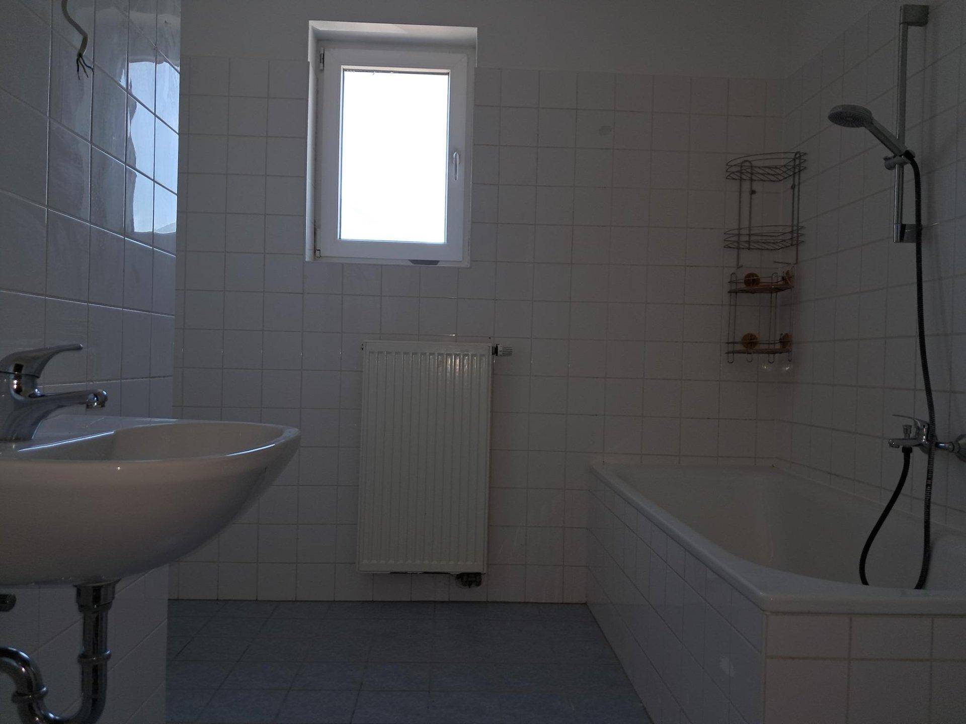 Badezimmer mit weißen Fliesen, Badewanne mit Duschfunktion, Waschbecken und einem Fenster.