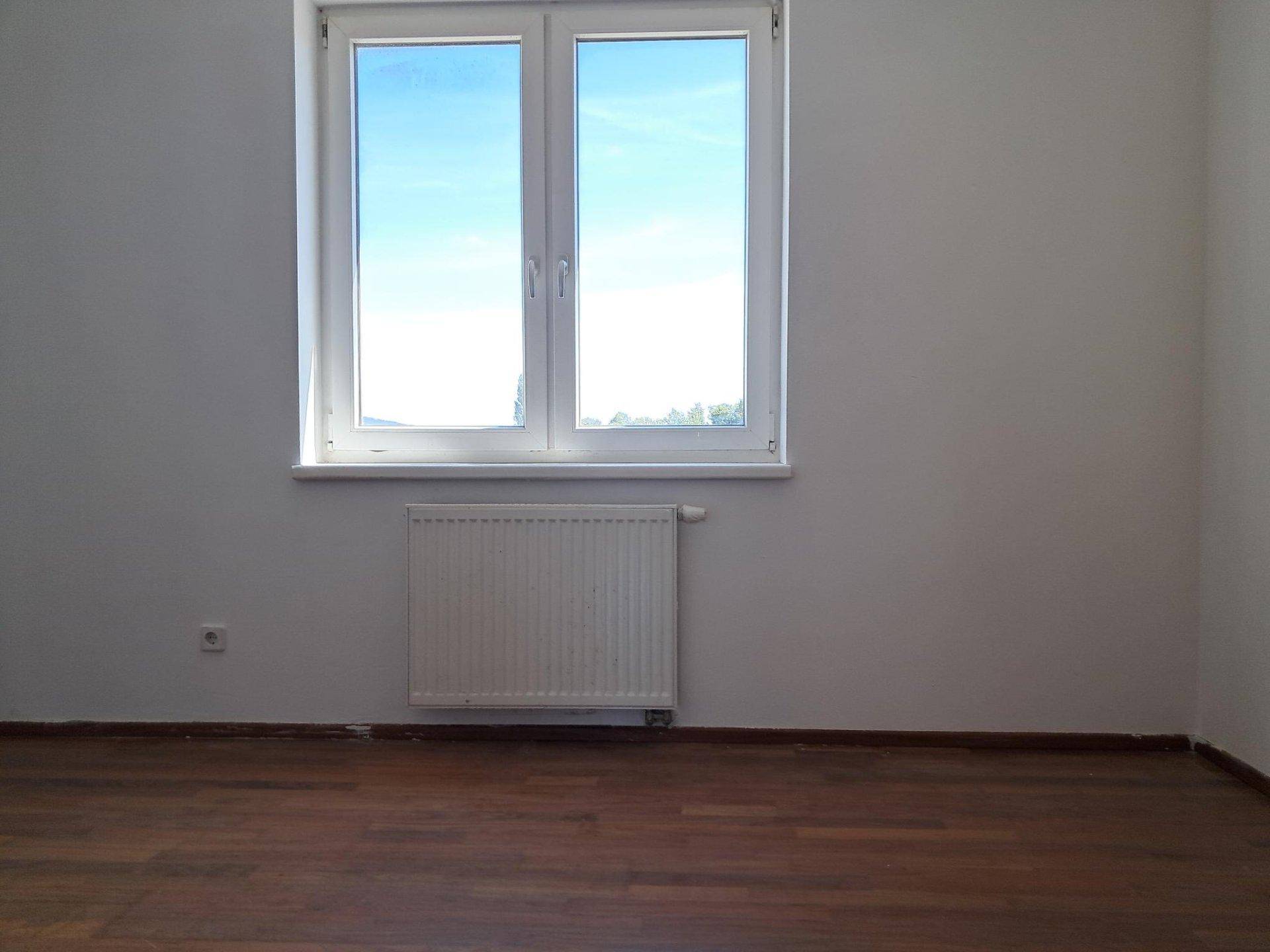 Helles, leeres Zimmer mit dunklem Holzboden, weißen Wänden und einem großen Fenster.