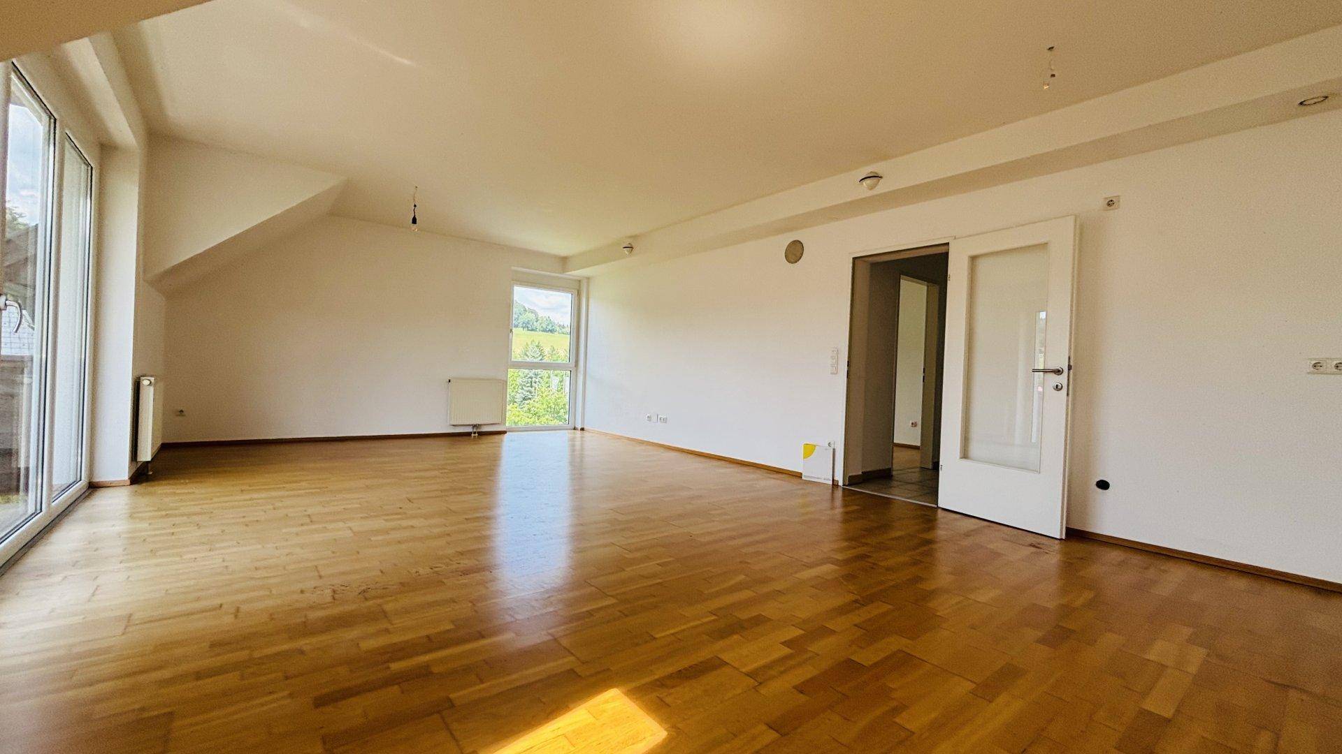 Geräumiges Wohnzimmer mit Parkettboden, großen Fenstern und Blick auf die grüne Umgebung.