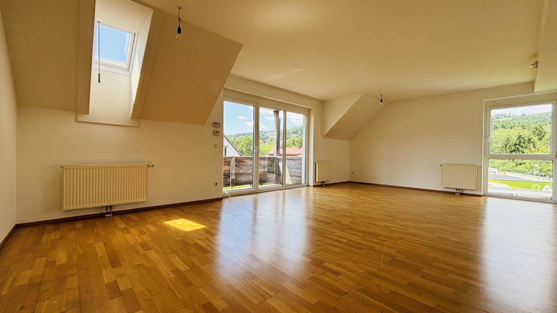 Helles, leeres Wohnzimmer mit Holzboden, Dachschrägen und Zugang zu einem Balkon mit Bergblick.