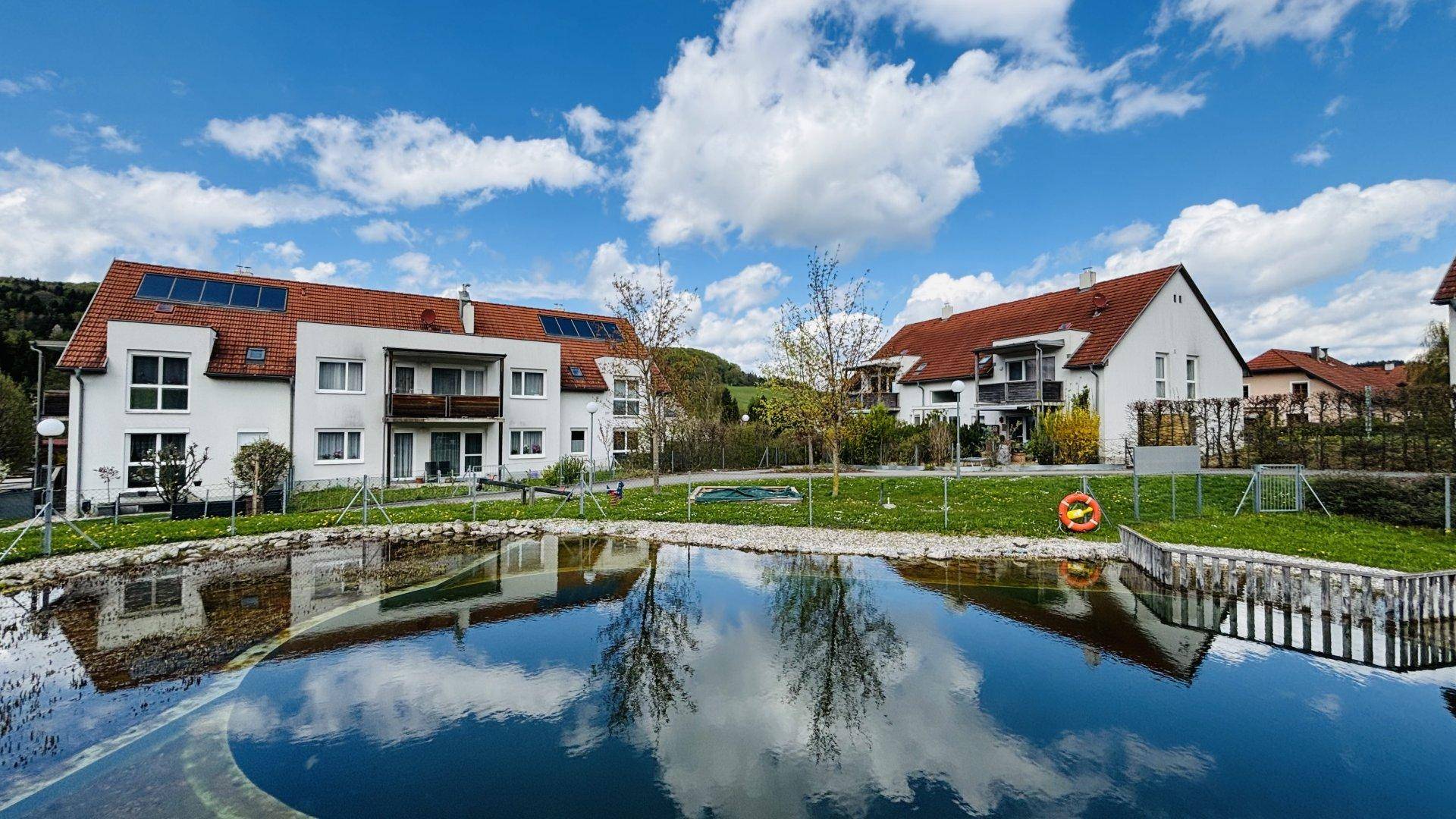 Moderne Wohnanlage mit mehreren Gebäuden, Solaranlagen und einem spiegelnden Teich unter blauem Himmel.