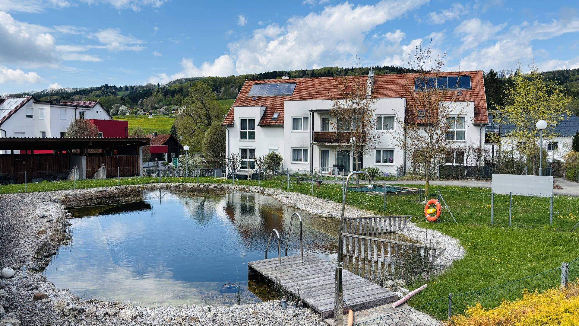 Wohngebäude mit Solarpaneelen auf dem Dach, umgeben von einer grünen Landschaft und einem Naturteich.