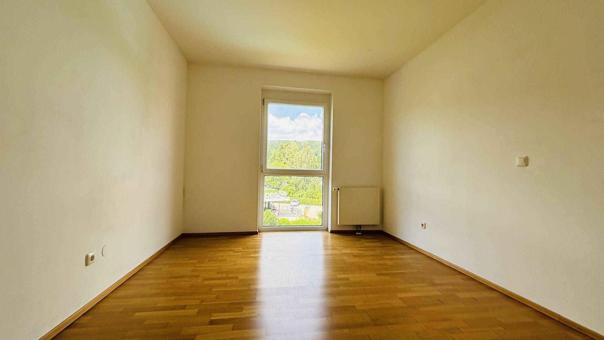 Leeres Schlafzimmer mit Parkettboden und einem hohen Fenster, das einen weiten Ausblick bietet.