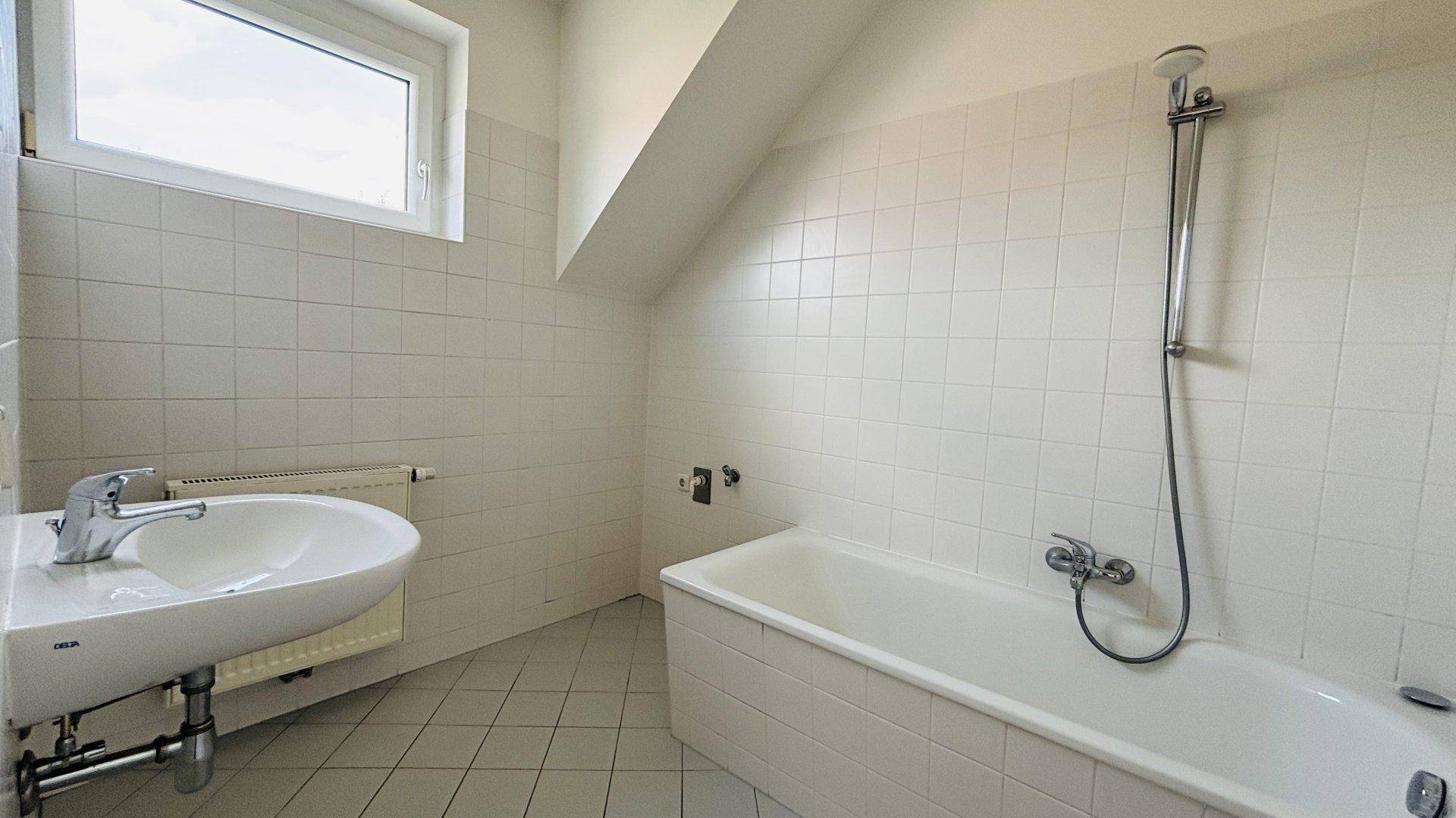 Badezimmer mit weißen Fliesen, Badewanne, Waschbecken und Dachschräge, gut erhalten.