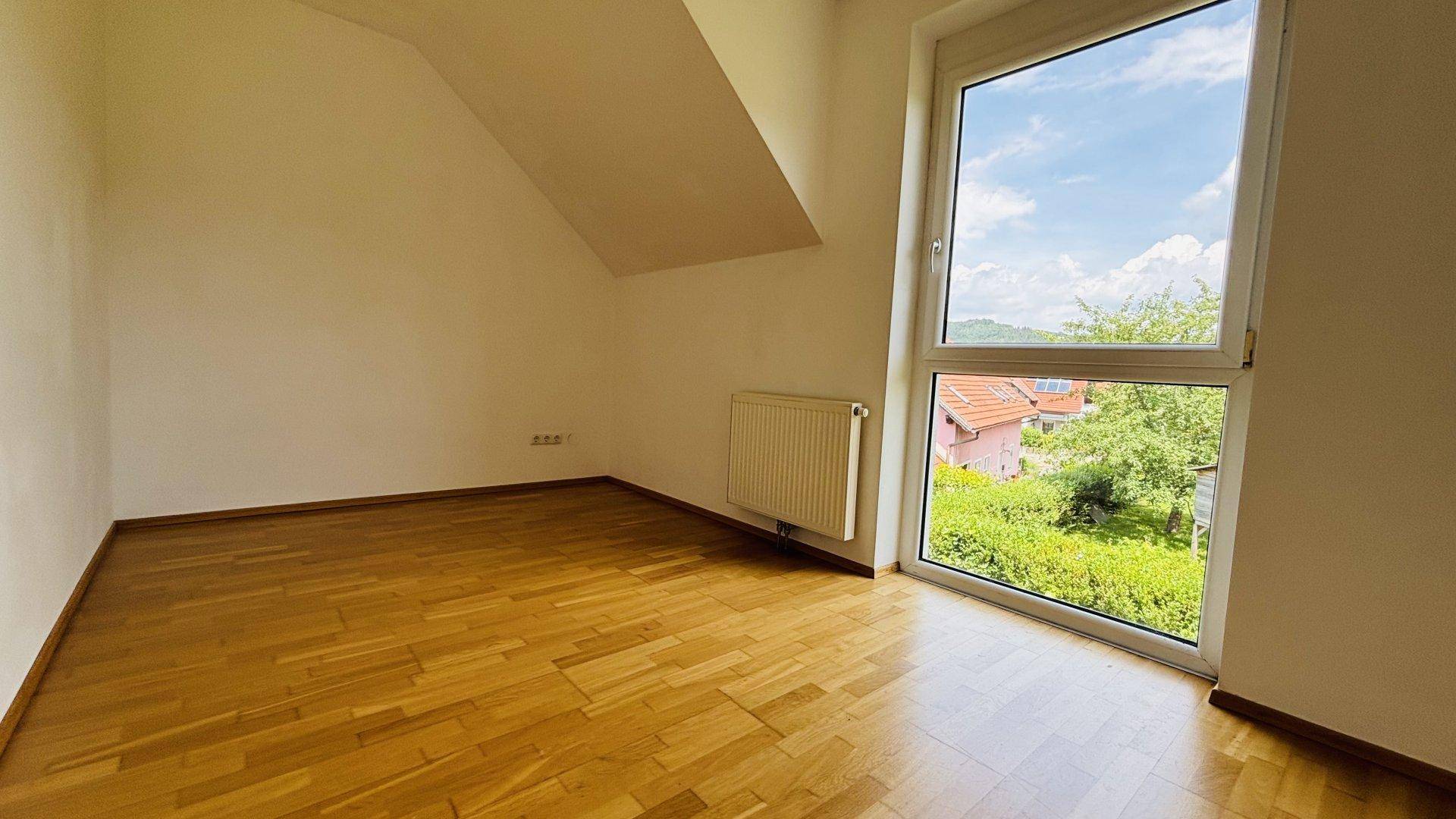 Helles Schlafzimmer mit Parkettboden, großem Fenster und Blick auf die grüne Umgebung.