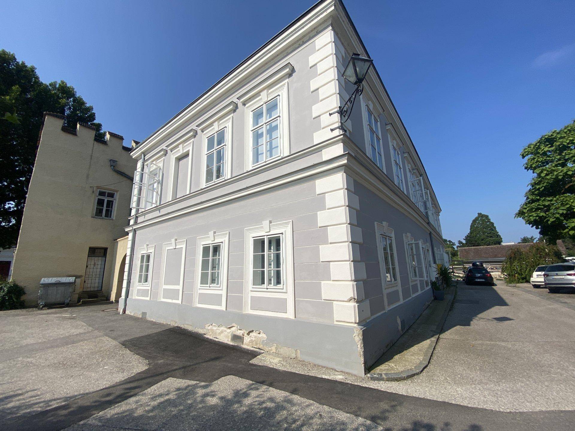 Eckansicht des historischen Gebäudes mit grauer Fassade, weißen Fensterläden und gepflastertem Bereich.
