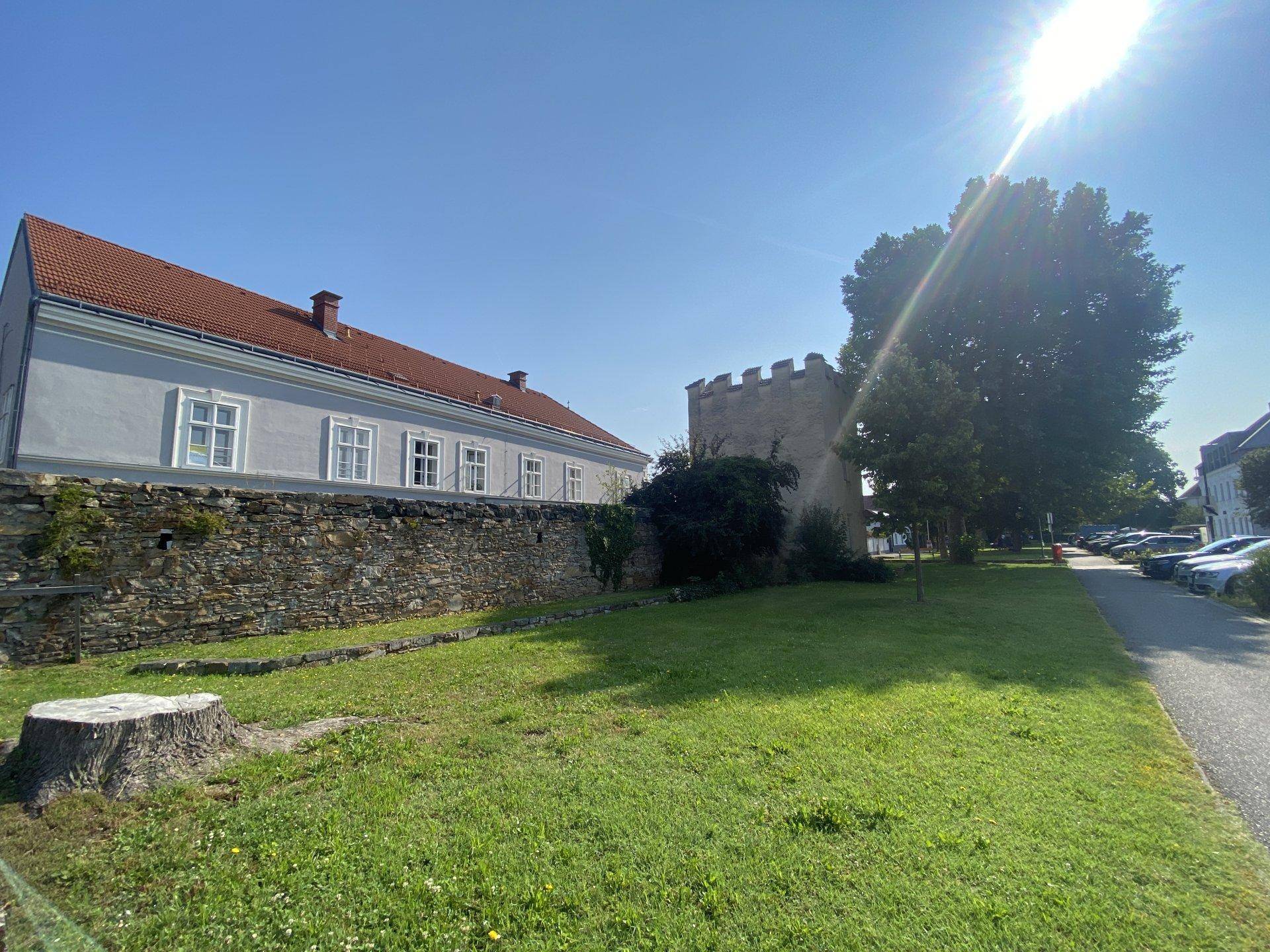Historisches Gebäude mit Steinmauer, grüner Wiese und Turm im Hintergrund bei Sonnenschein.
