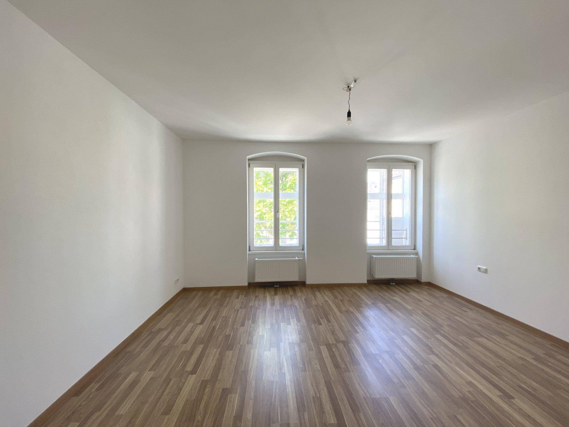 Geräumiger, leerer Raum mit Laminatboden, weißen Wänden und zwei Fenstern.