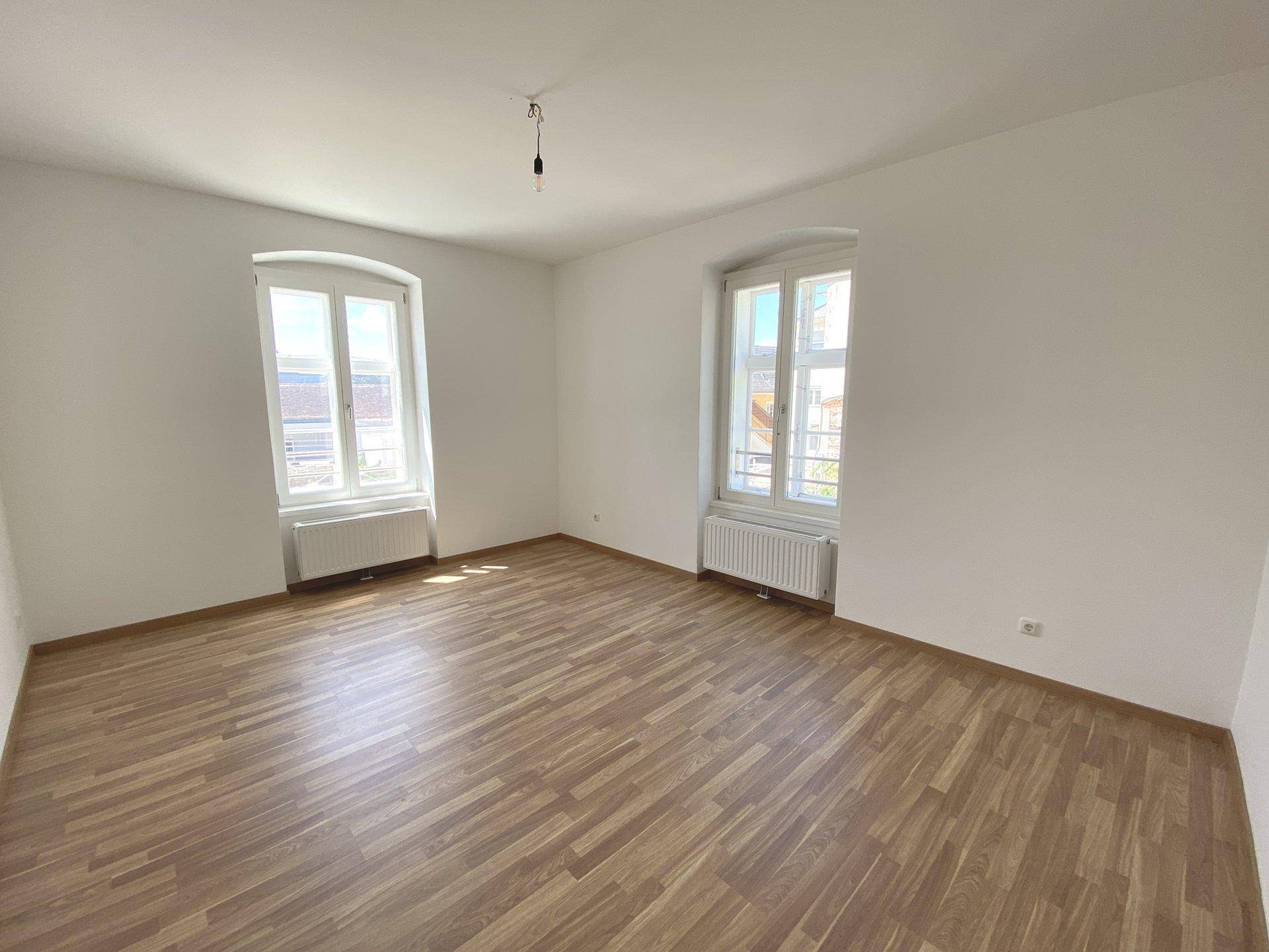 Heller Wohnraum mit Laminatboden, weißen Wänden und zwei großen Fenstern.