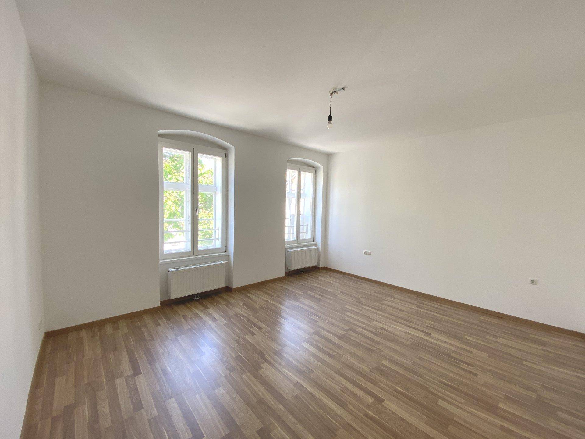 Lichtdurchfluteter Raum mit Laminatboden, weißen Wänden und zwei Fenstern mit Ausblick.