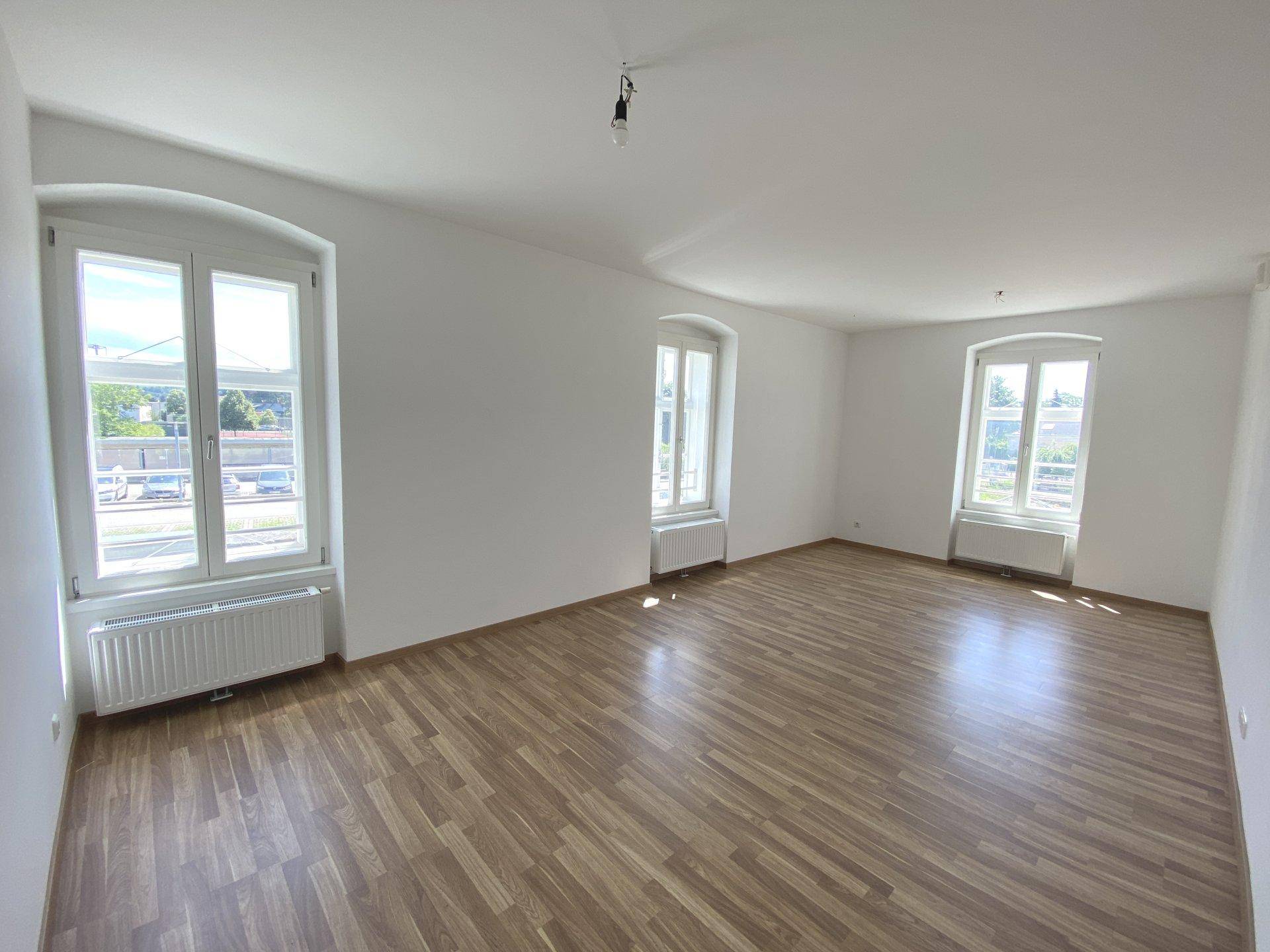 Großer, heller Wohnraum mit Laminatboden, weißen Wänden und drei Fenstern.